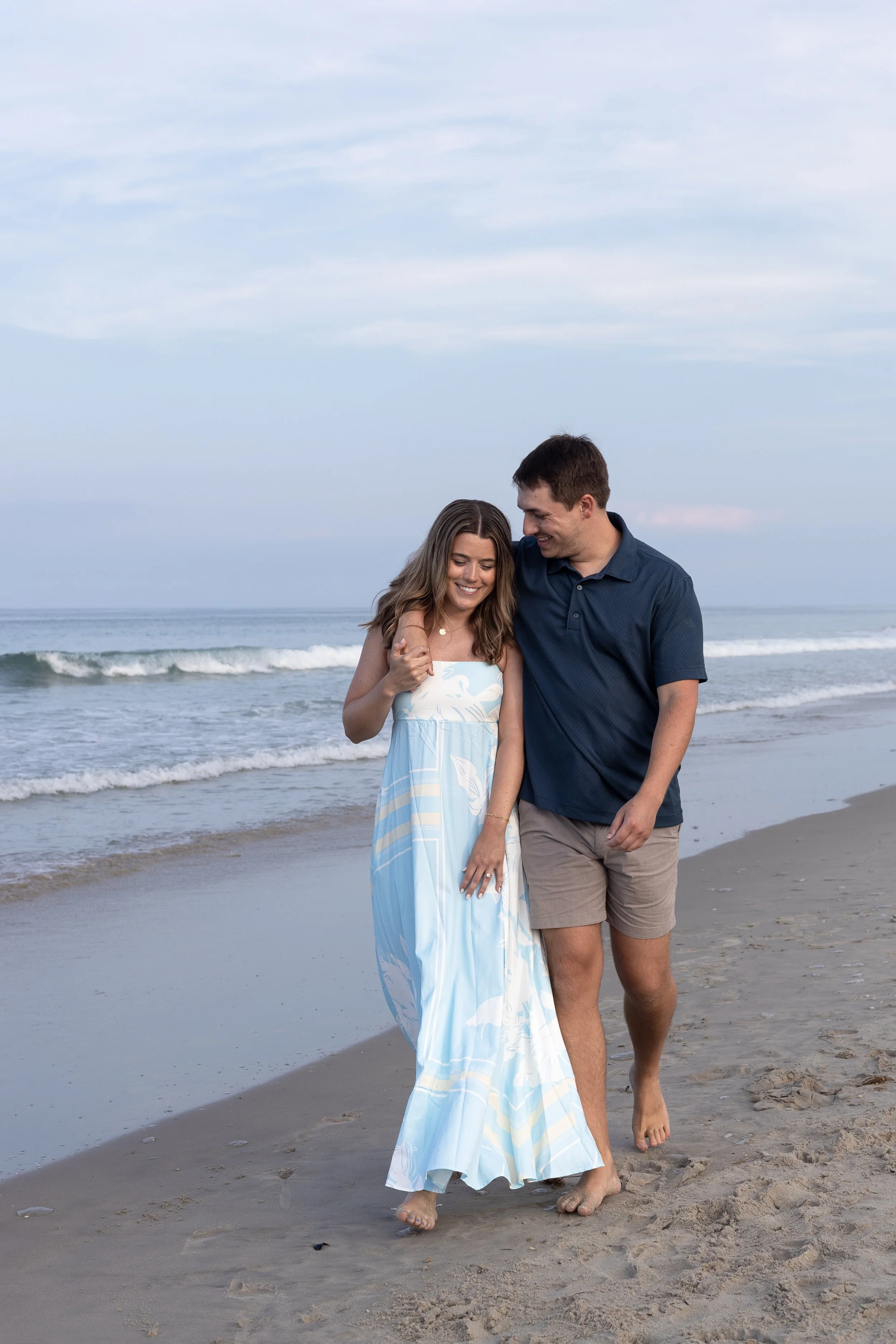 A young couple walking on the beach with the ocean and sky in the background, smiling and enjoying each other's company.