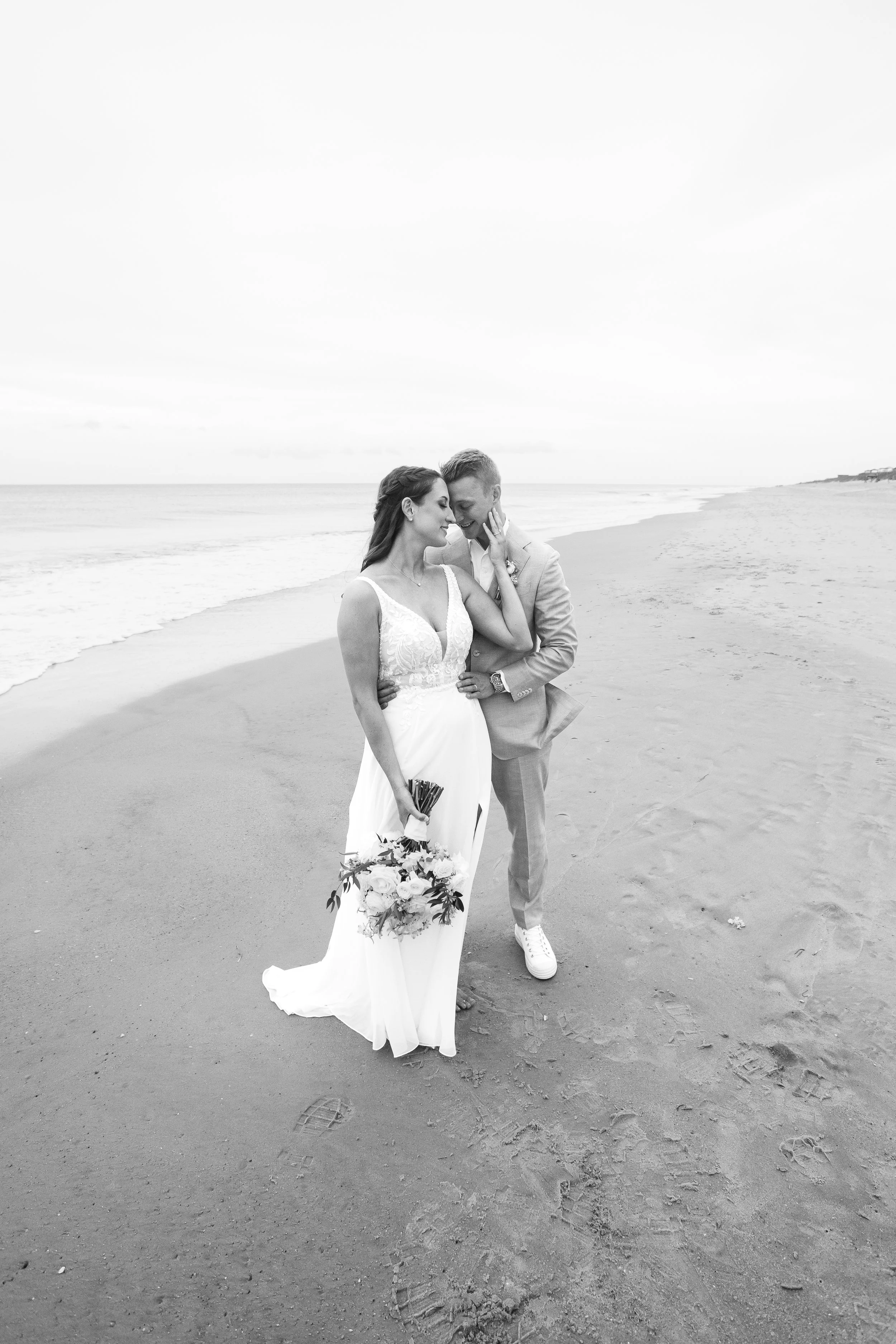 Black and white photo of a bride and groom standing together on the beach, close together, with their foreheads touching. The bride is holding a bouquet of flowers, and the groom is dressed in a light-colored suit, wearing sneakers. The ocean and sho