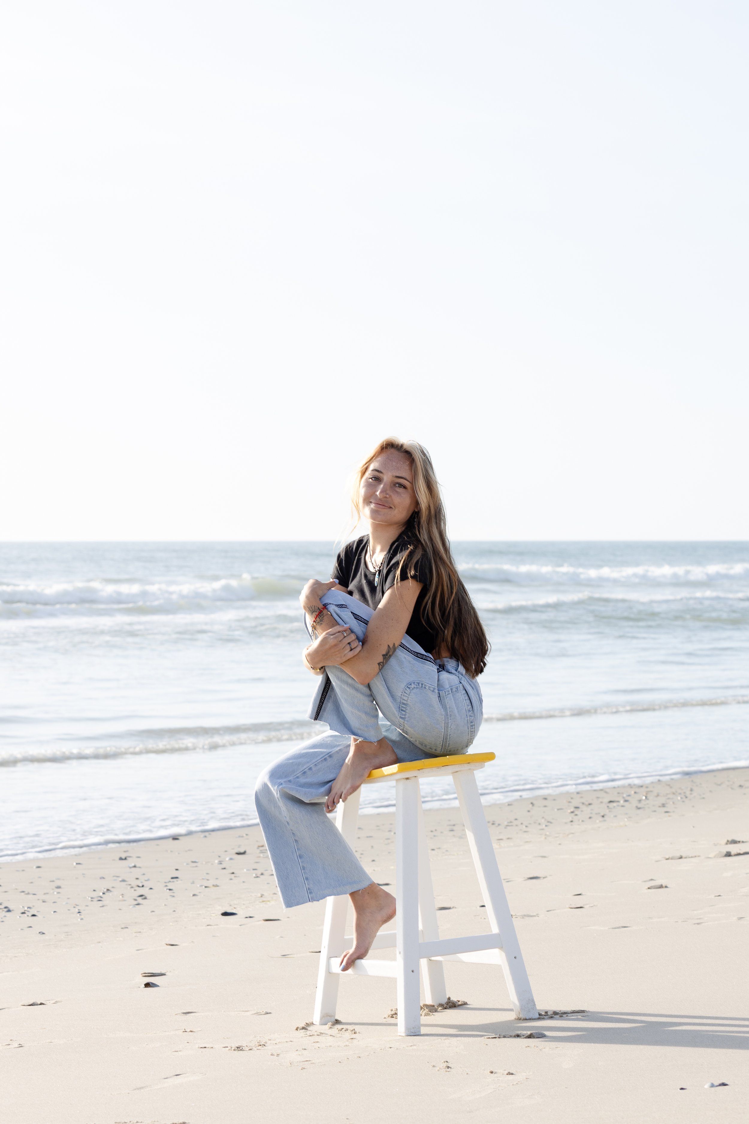 A young woman with long hair sitting on a yellow and white stool on a sandy beach near the ocean, smiling at the camera.