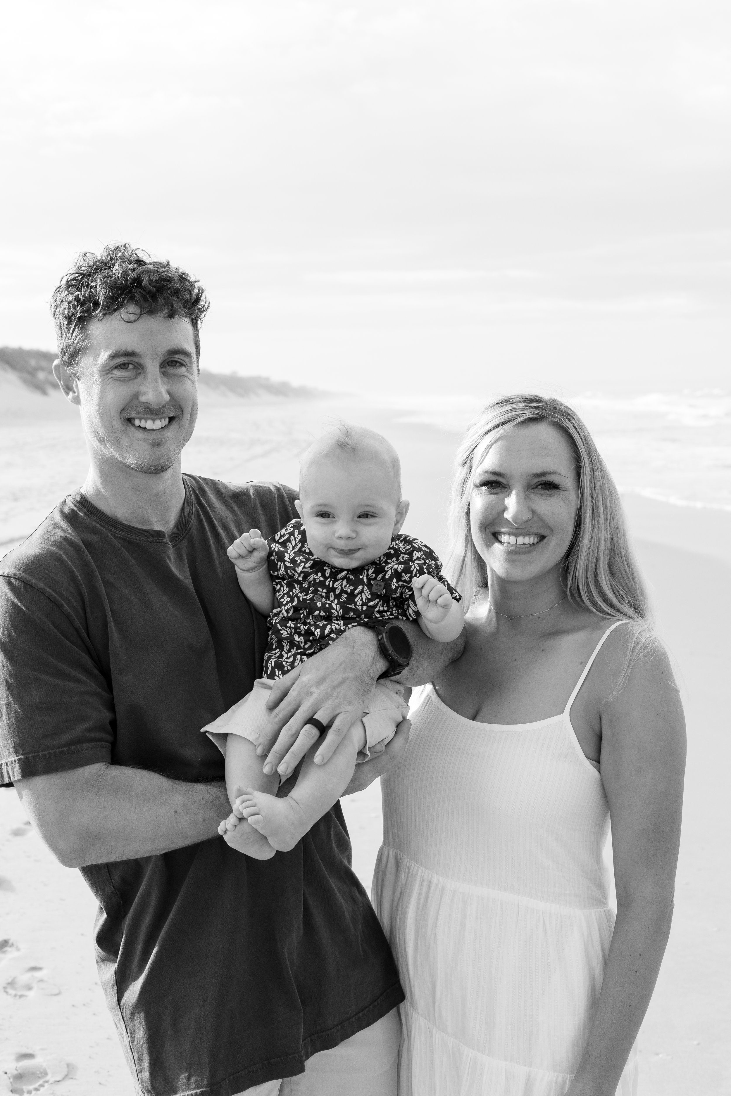 A black and white photo of a smiling family of three on a beach; a man holding a baby girl, and a woman standing beside them, all facing the camera.