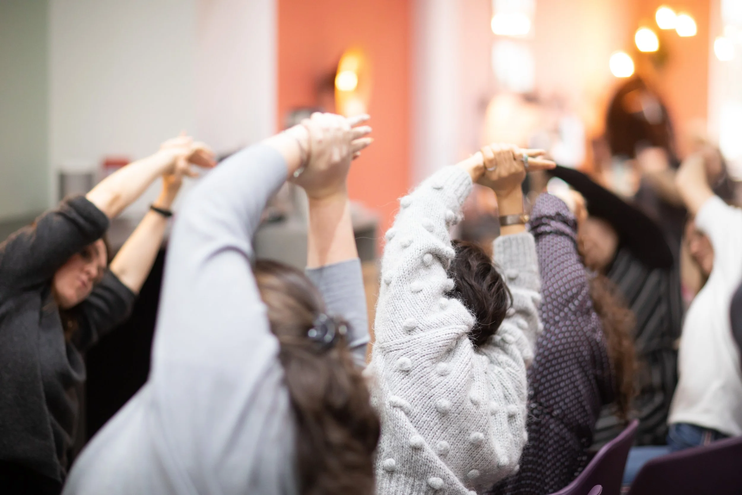 A woman leading a stretch or yoga session for a group of seated people in an indoor setting with natural light, plants, and a coffee station.