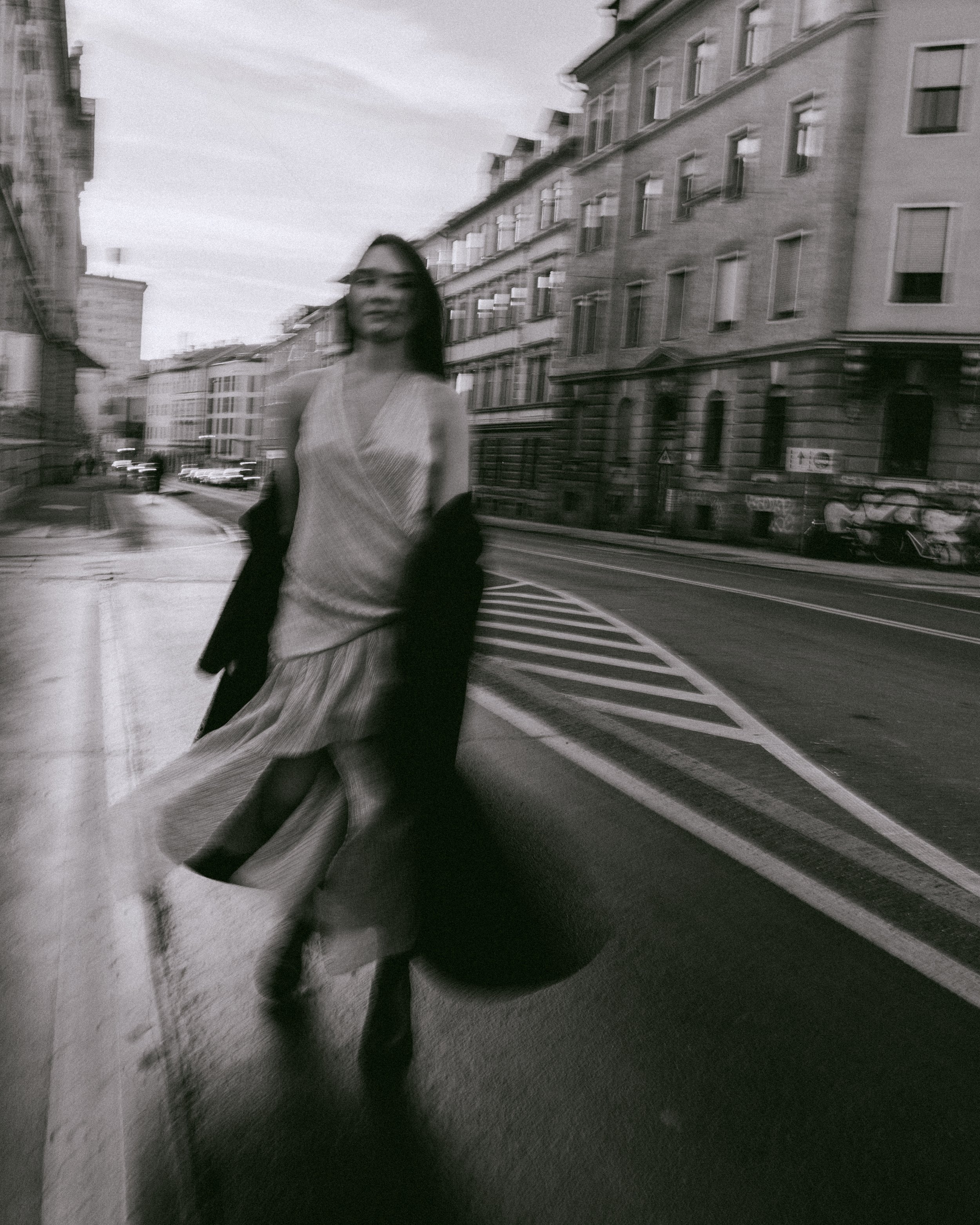 Schwarz-weiß-Foto einer Frau in einem Kleid, die auf einer Stadtstraße läuft, mit Gebäuden im Hintergrund.