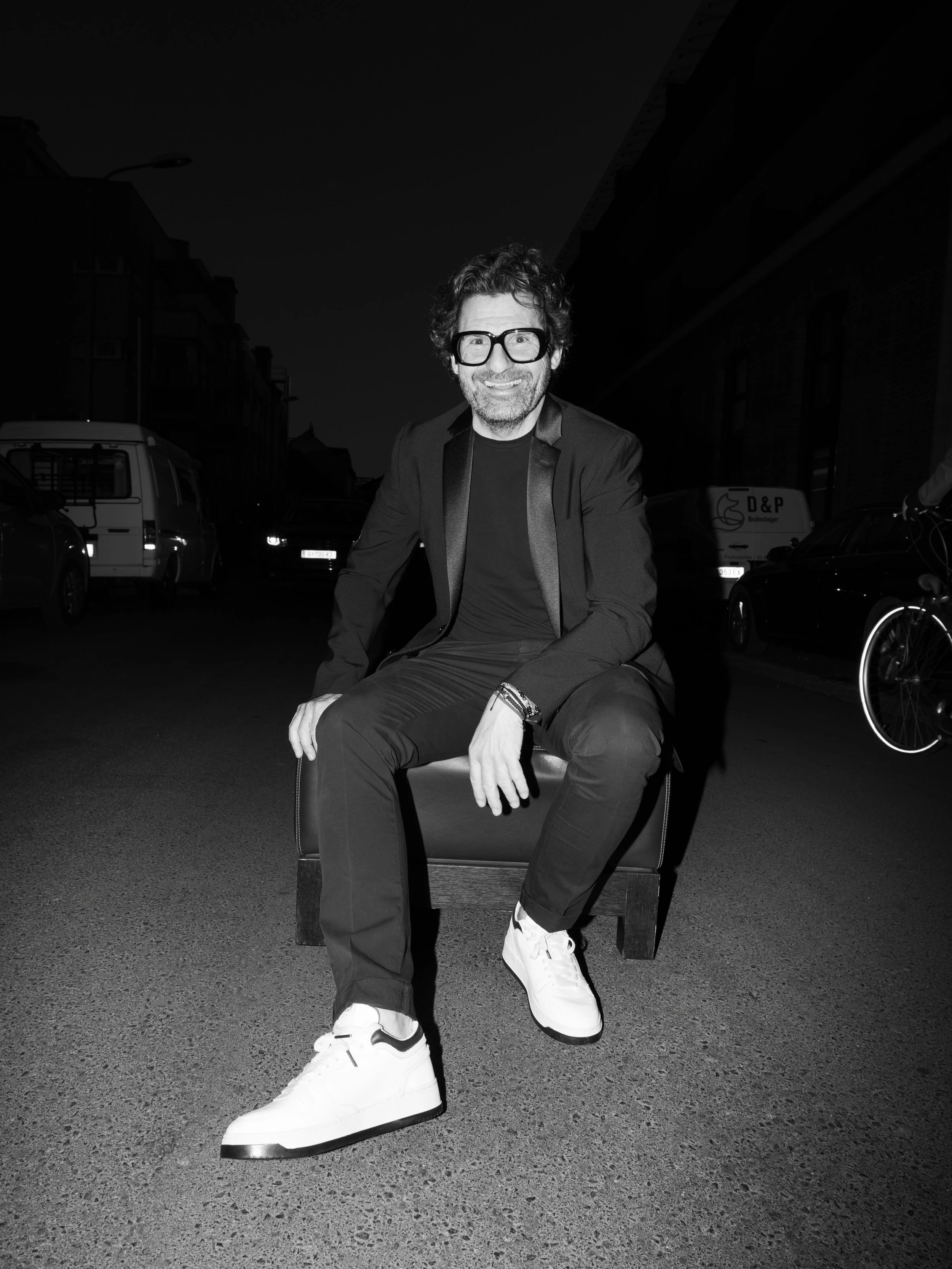 Ein Mann mit großen Brillen sitzt lächelnd auf einem Stuhl auf einer Straße bei Nacht.