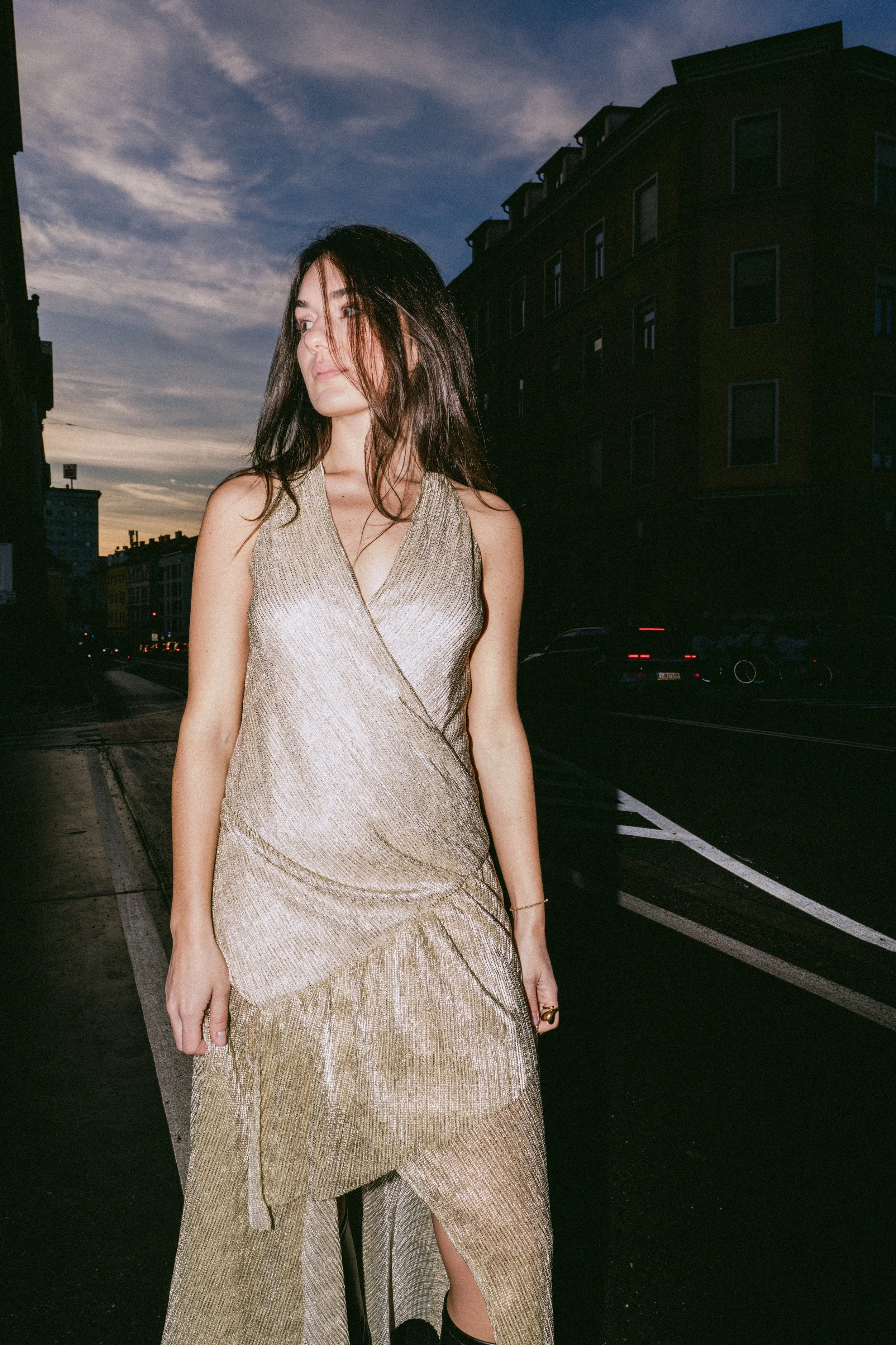 Junge Frau in funkelndem goldfarbenem Kleid steht auf einer dunklen StraÃe bei Sonnenuntergang.