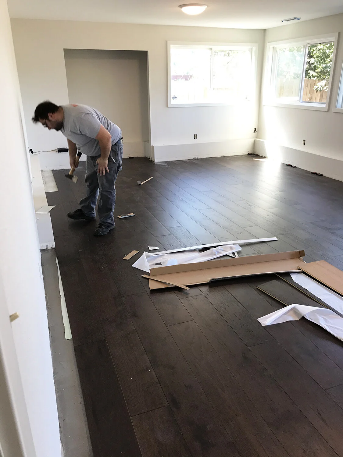 A man installing dark hardwood flooring in a bright, empty living room with large windows.