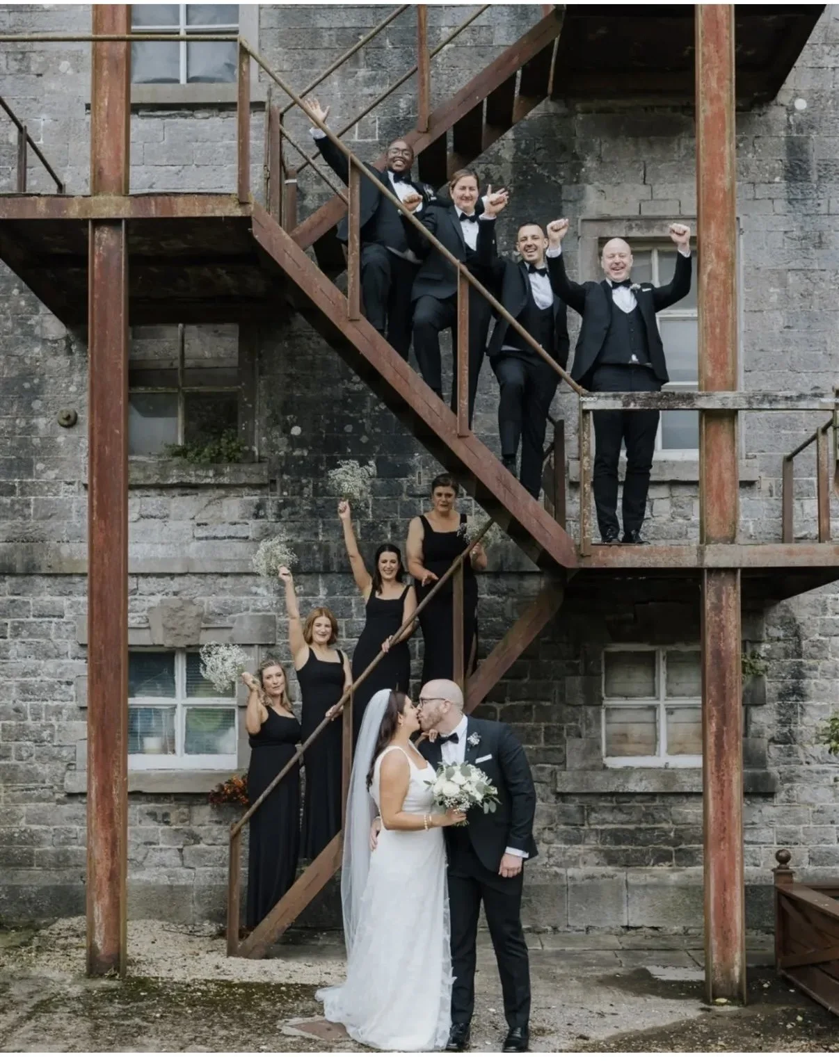 Wedding couple kissing in front of a rustic stone building with bridesmaids and groomsmen on a staircase celebrating.
