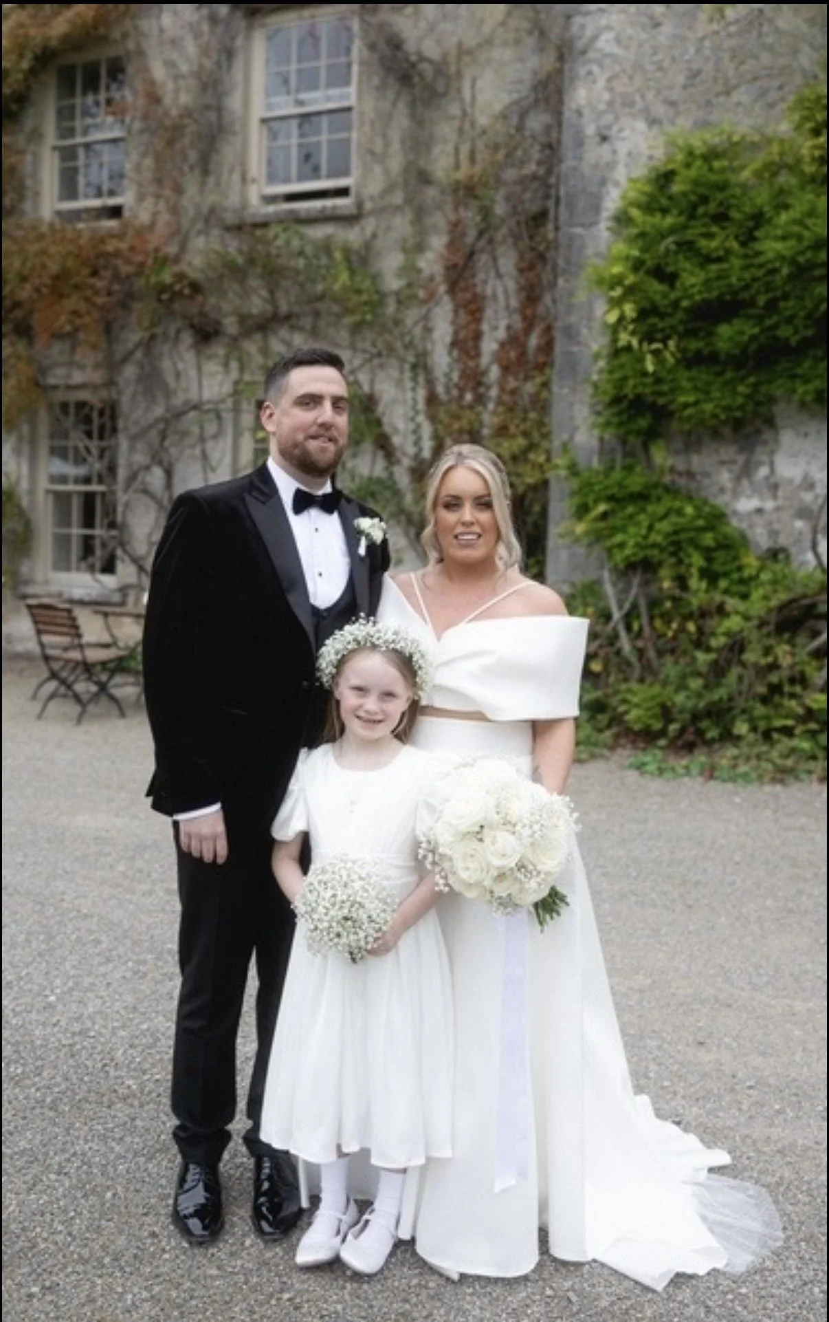 A wedding photo featuring a man in a black tuxedo, a woman in a white wedding dress, and a young girl in a white dress holding a bouquet of white flowers. They are standing outdoors in front of a building with ivy-covered stone walls.