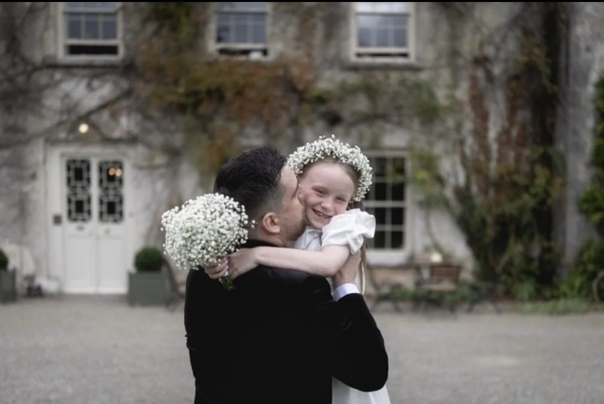 A man in a black suit holding a smiling young girl with a flower crown, both holding bouquets of white flowers, outside a stone house with ivy-covered walls.