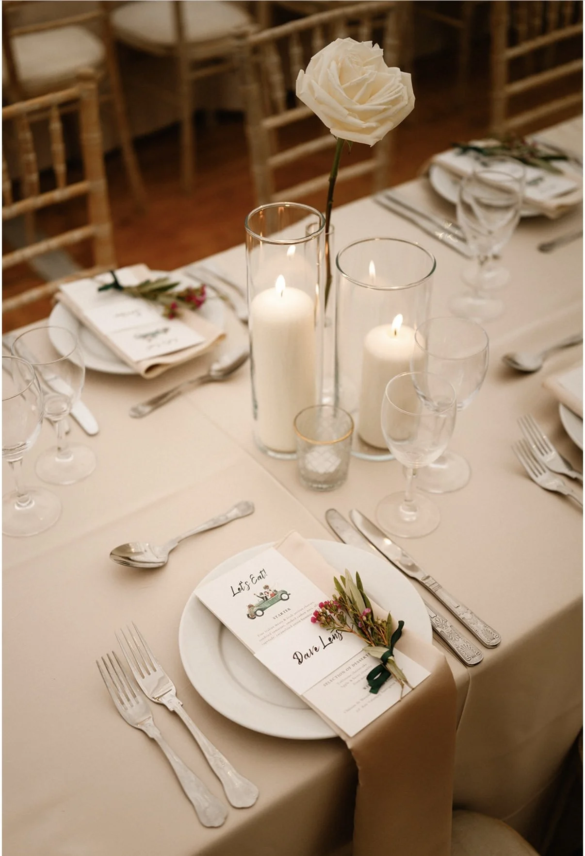 Elegant wedding table setting with white place settings, silverware, wine glasses, candles, and a white rose centerpiece with greenery.