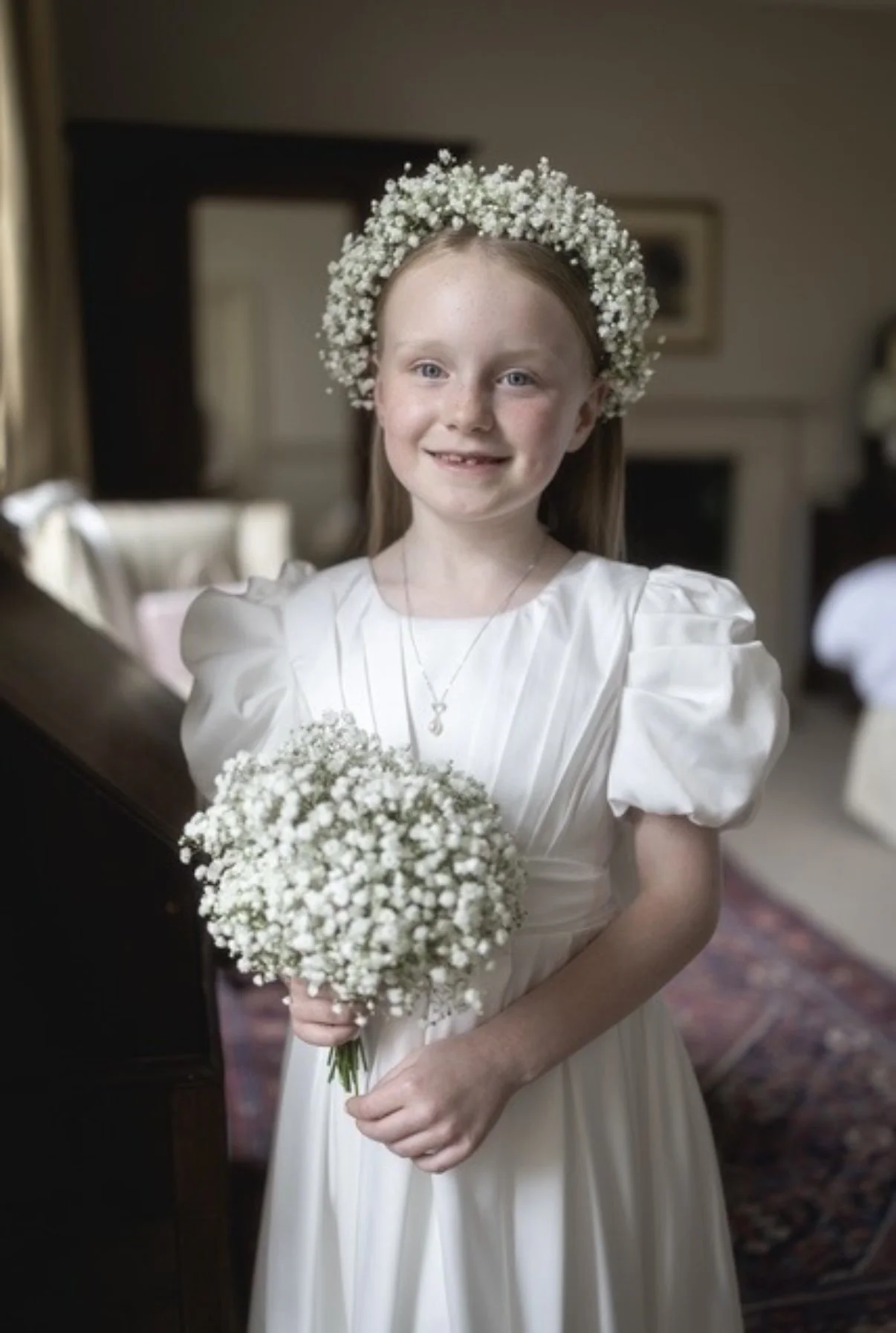 A young girl with red hair, wearing a white dress and a floral crown made of small white flowers, holding a bouquet of white flowers, standing indoors with a blurred background.