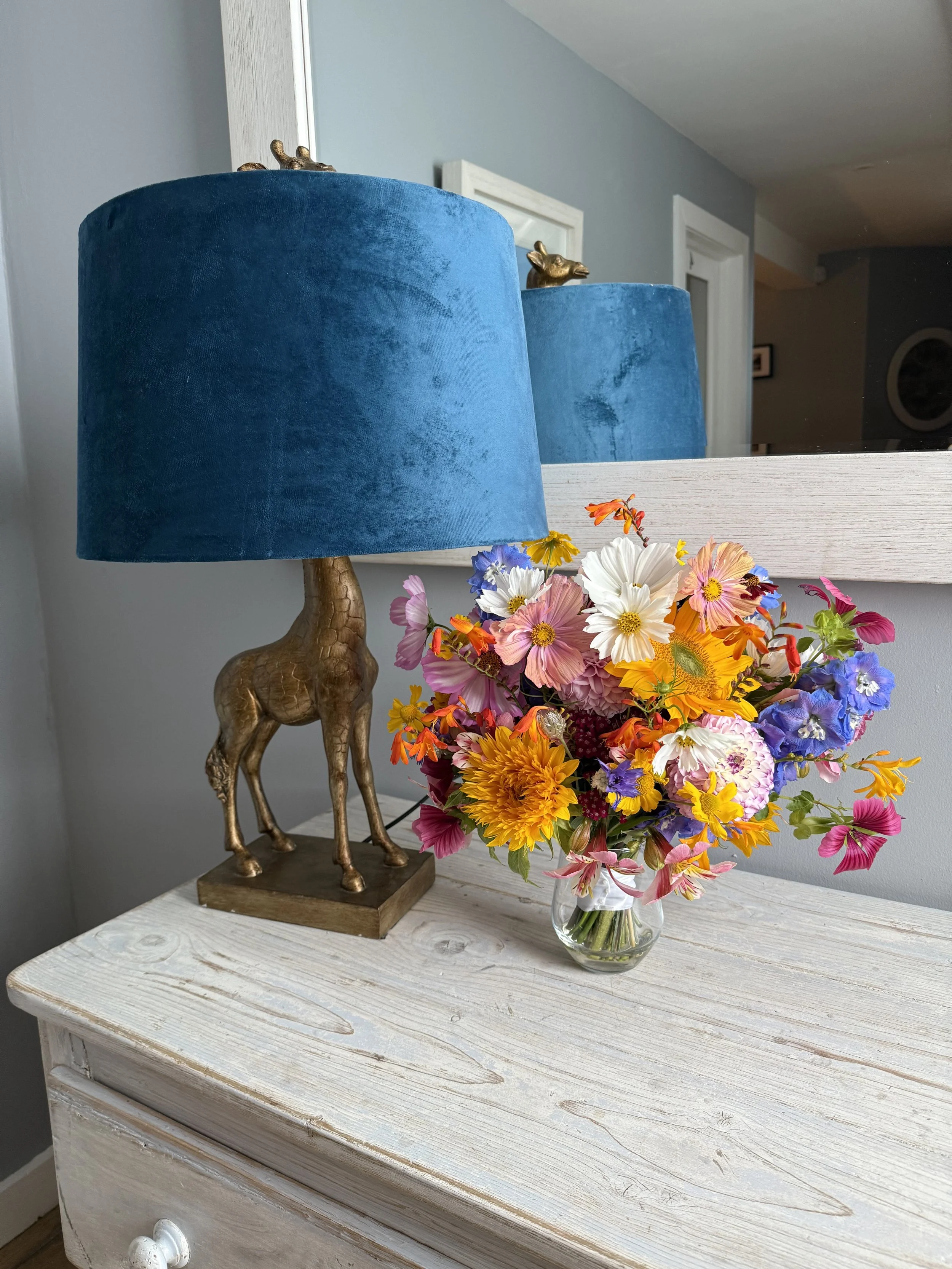 A table lamp with a giraffe-shaped base and a blue lampshade, placed on a white wooden table next to a colorful bouquet of flowers in a glass vase.