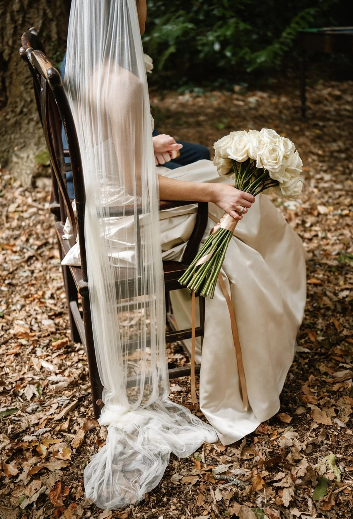 A bride sitting on a wooden chair outdoors in a wooded area, holding a bouquet of white roses, with a sheer veil draped over the chair and some leaf litter on the ground.