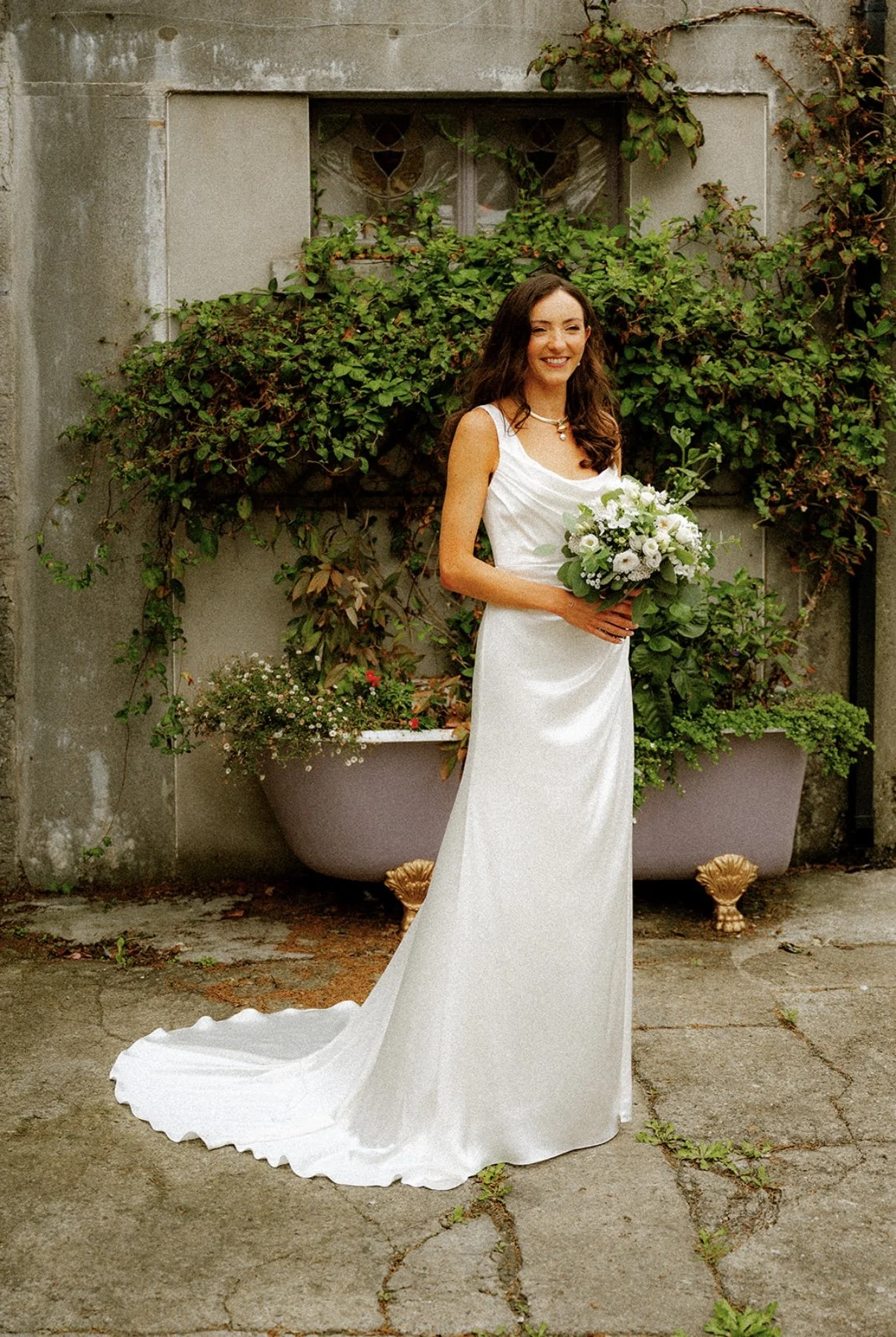 A woman in a white wedding dress holding a bouquet of white and green flowers, standing outdoors in front of a concrete wall and large potted plants with foliage.