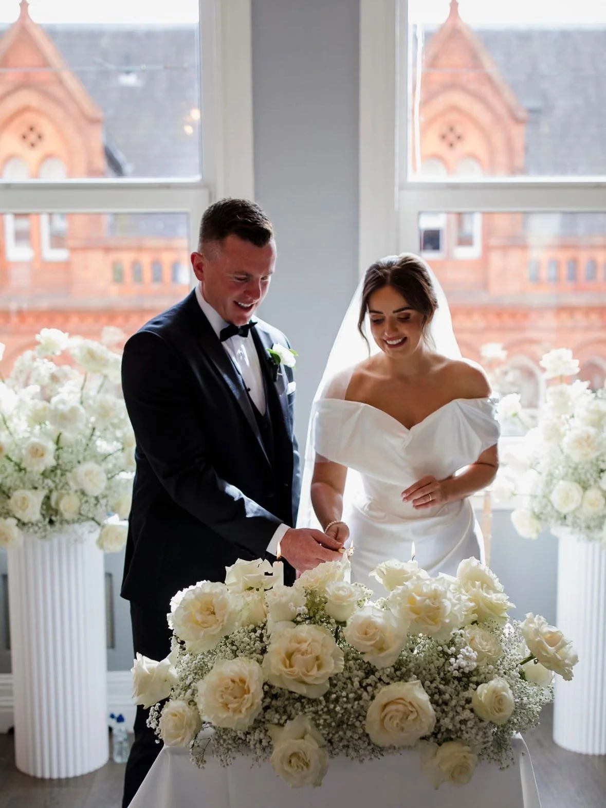 Bride and groom in wedding attire lighting a candle on a table decorated with white roses and baby's breath during a wedding ceremony, with large windows and a historic building in the background.