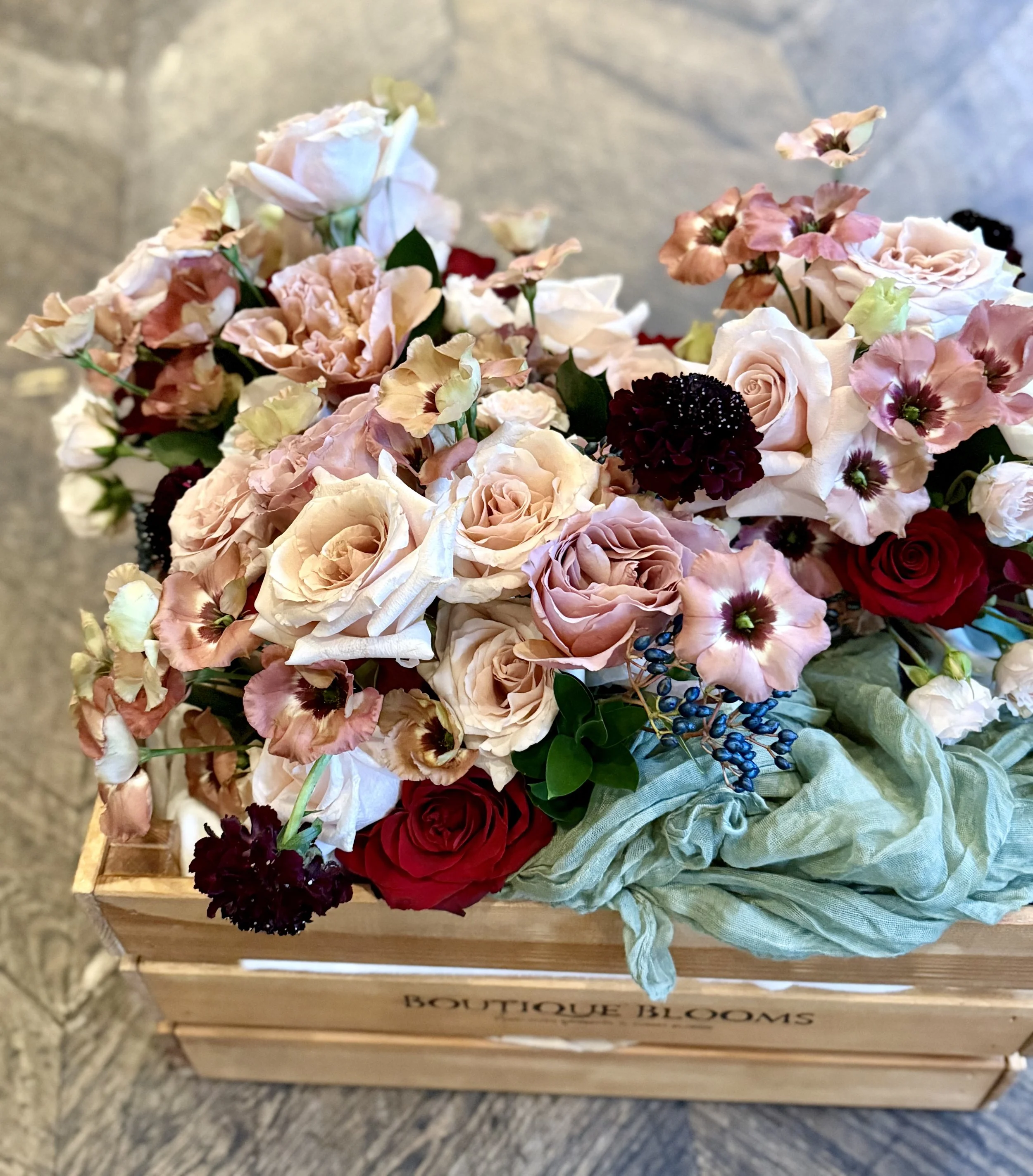 A wooden crate filled with a variety of pink, burgundy, and dark red flowers with green foliage and a green cloth around the base.
