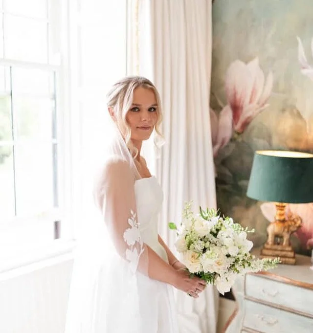 A bride standing inside with a bouquet of white flowers, wearing a white wedding dress, near a window and a floral mural on the wall.