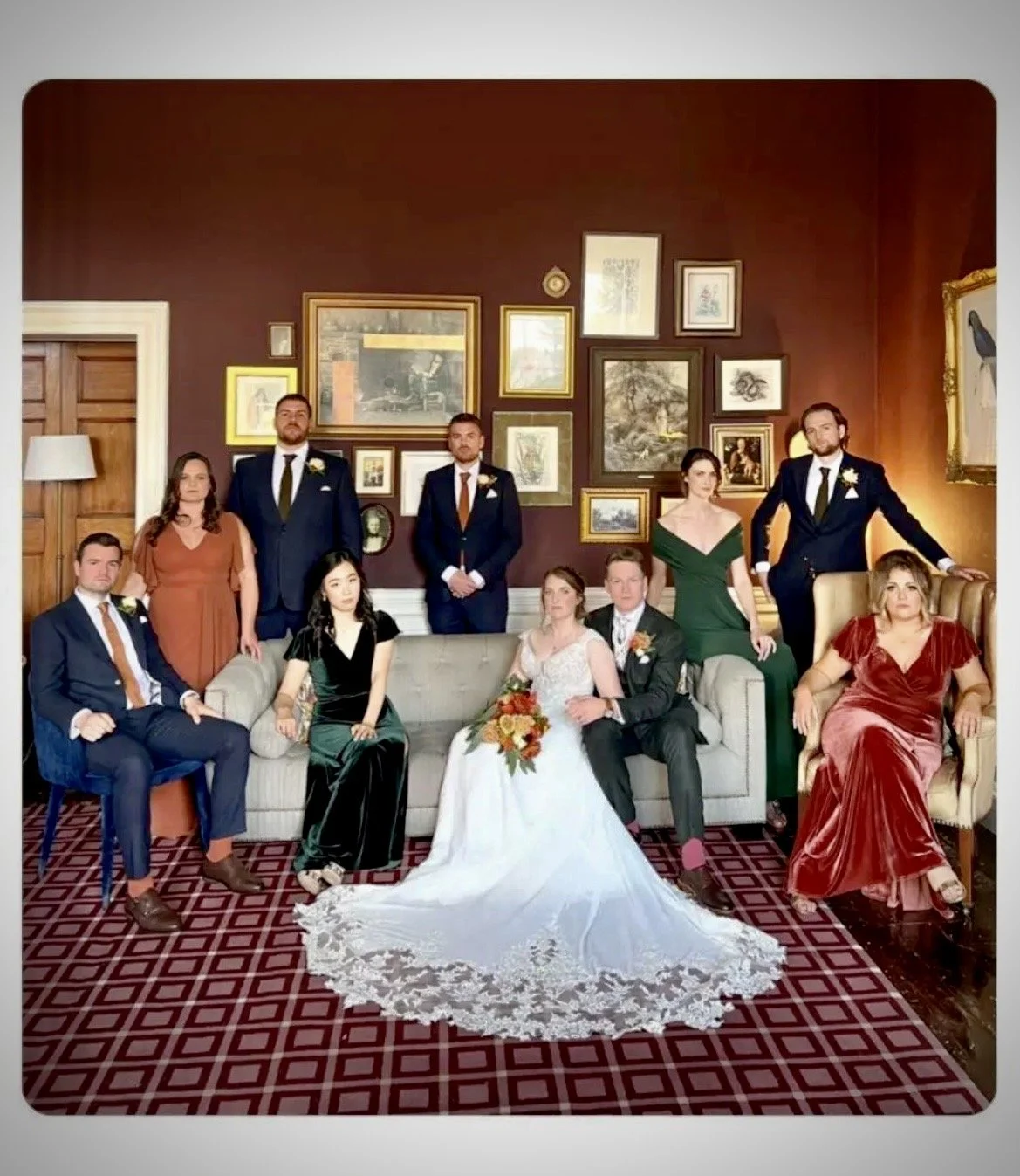 A wedding party with 11 people posing in a decorated room, with a bride and groom seated in the center, surrounded by bridesmaids, groomsmen, and other guests.