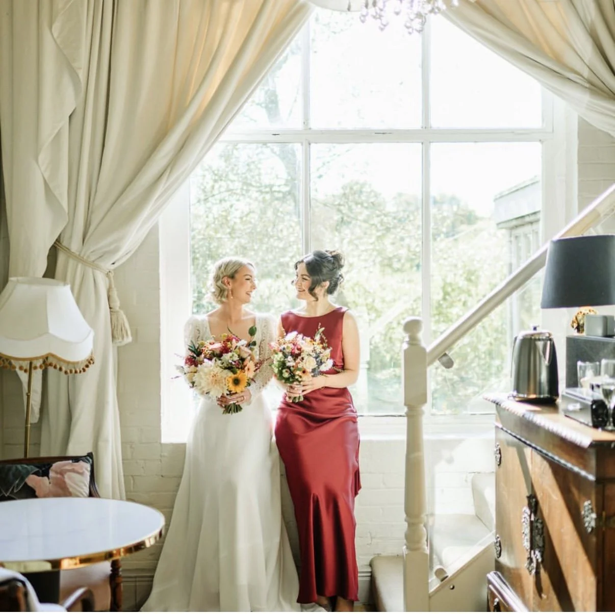Two women, one in a white wedding dress and the other in a red dress, standing by a large window with flowing curtains, holding colorful bouquets and smiling at each other in a bright, cozy room.