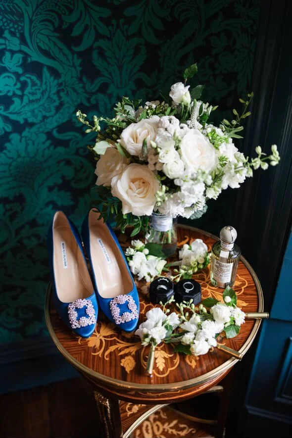 A round wooden table with intricate inlay designs holding a bouquet of white roses and greenery, a pair of blue high heels with sparkling embellishments, black camera, perfume bottle, and white flowers.