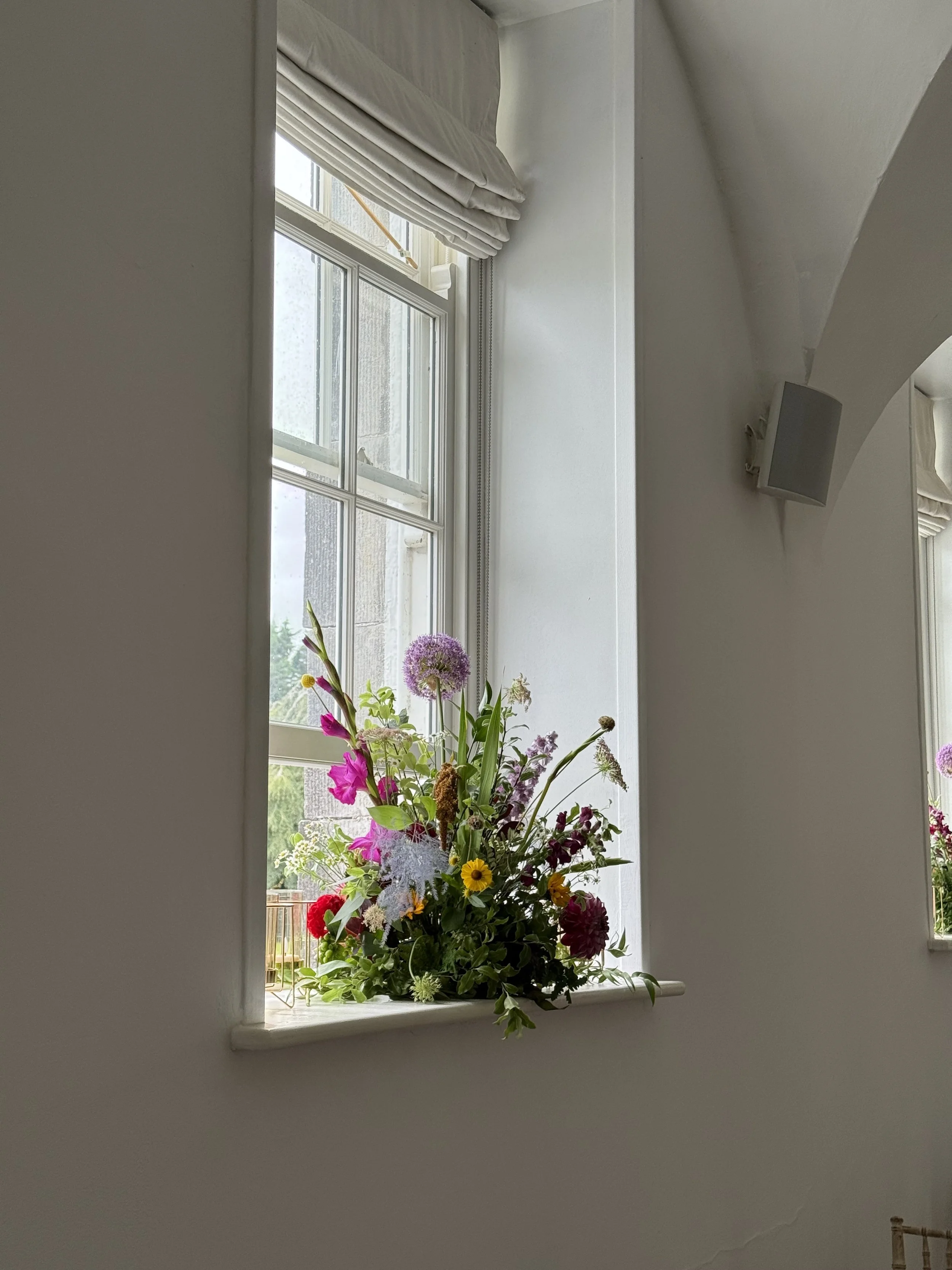 A windowsill with a colorful bouquet of various flowers, with a window in the background showing part of a building and some outdoor greenery.