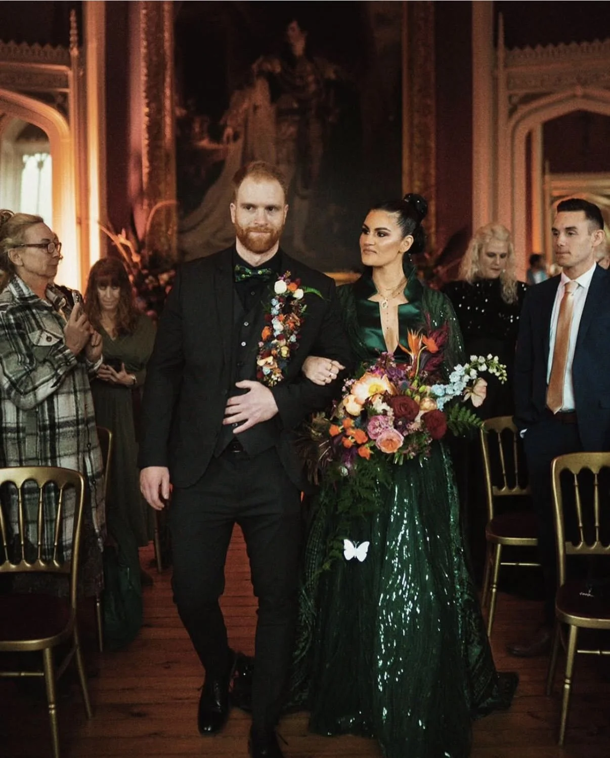 A man and woman walking arm in arm at a formal event, with the man wearing a black tuxedo with a floral boutonniere and the woman wearing a dark green dress with a vibrant bouquet of flowers, in a decorated ornate room.