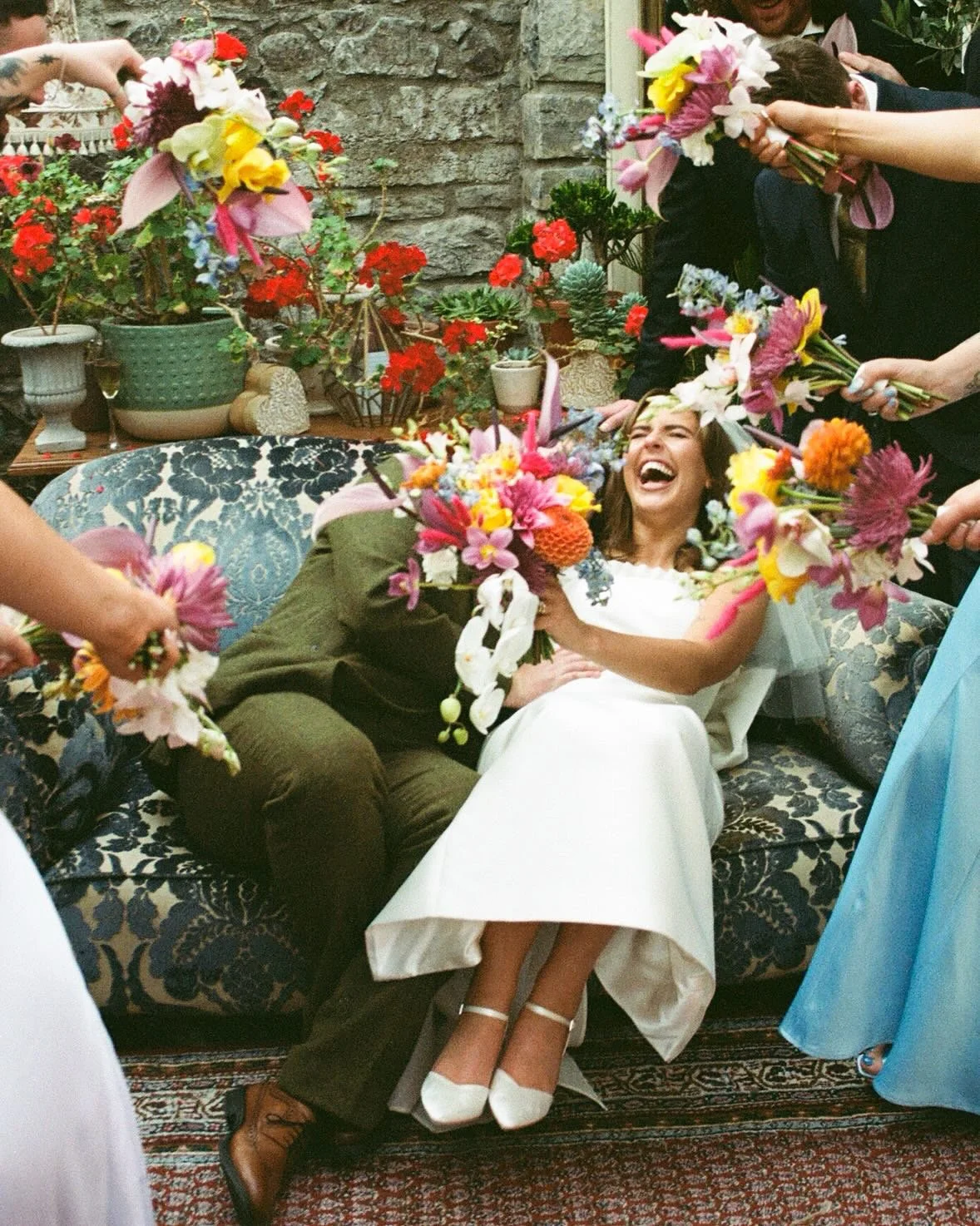 A woman in a white dress and a man in brown and black clothing are sitting on a vintage floral sofa, laughing as several people shower them with colorful flower bouquets during a celebration.