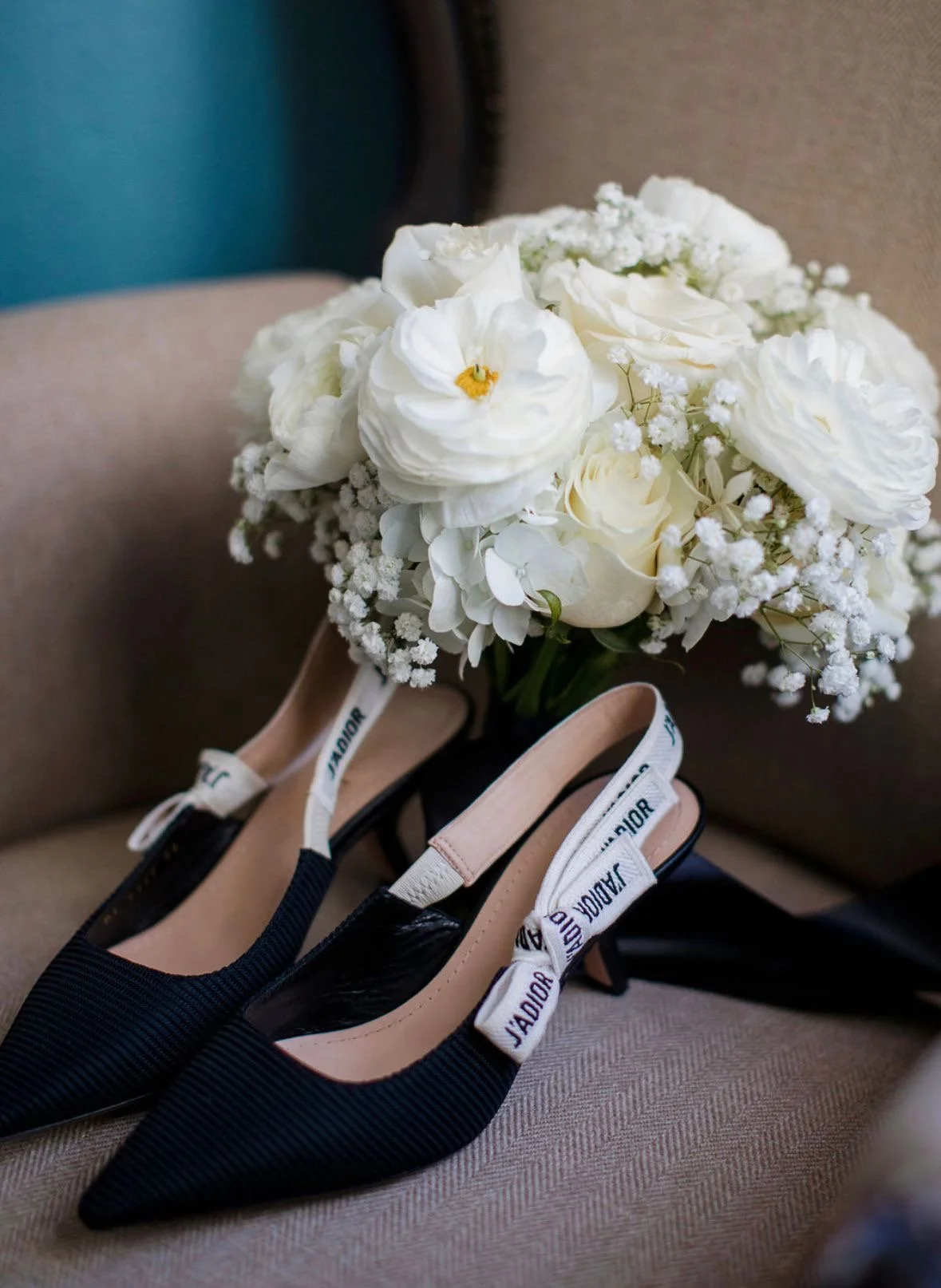 A bouquet of white flowers resting on a beige armchair with a pair of black and tan high-heeled shoes with white straps marked 'JADIOR' placed in front of it.