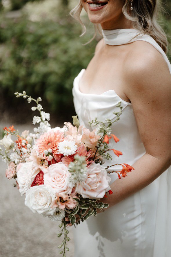A bride in a white satin dress holding a bouquet of pink, white, and peach flowers, with greenery, outdoor setting.