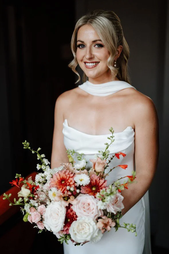 A woman in a white wedding dress holding a large bouquet of pink, orange, and white flowers, standing indoors and smiling at the camera.