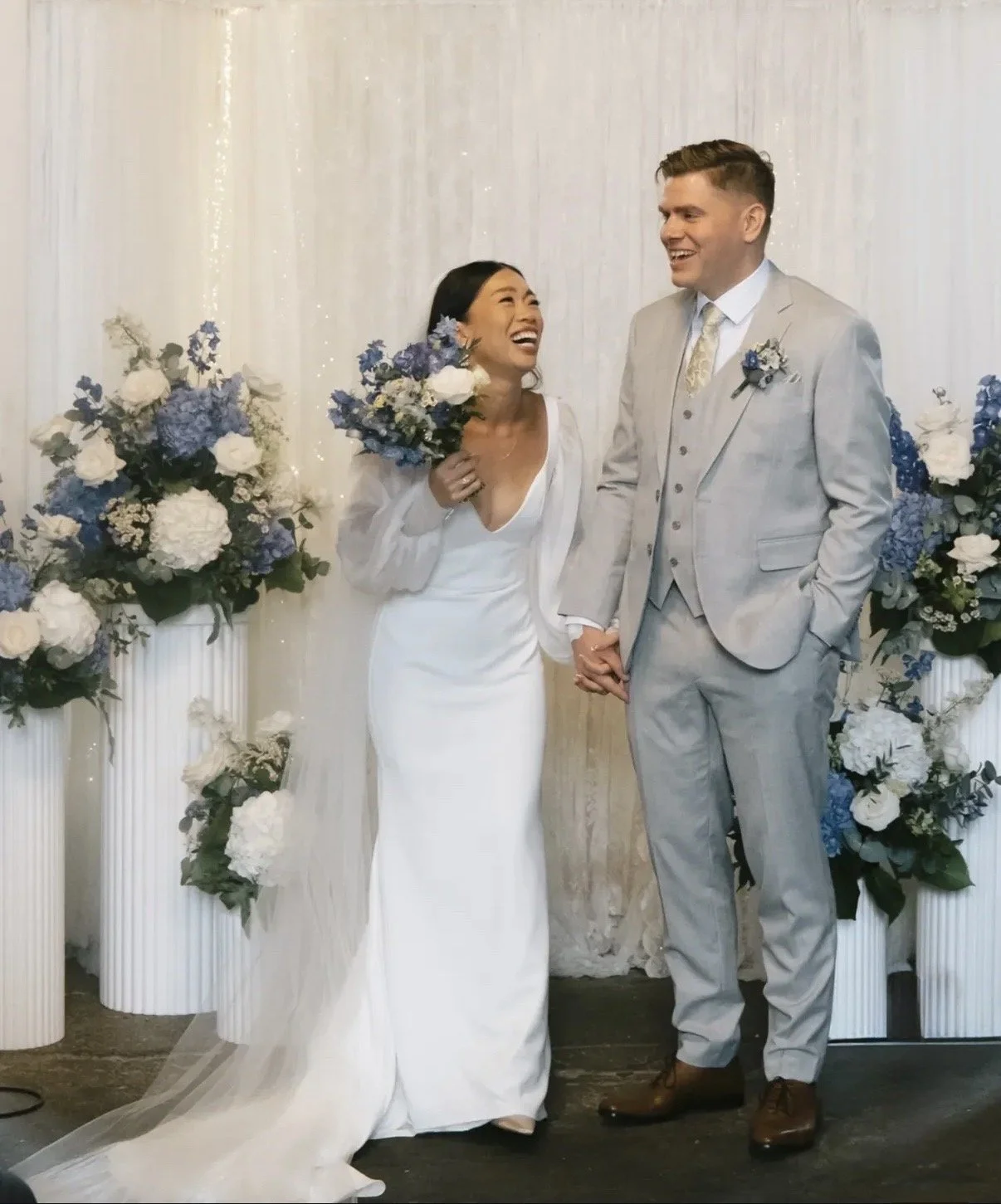 A bride and groom holding hands during their wedding ceremony, surrounded by flowers in white and shades of blue.