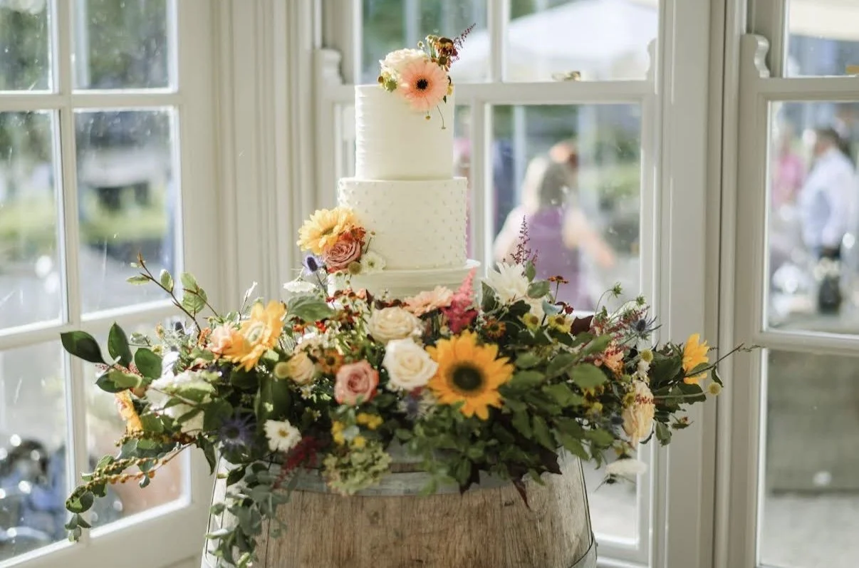 A wedding cake with two tiers decorated with flowers, placed on a wooden barrel surrounded by an autum  colorful floral arrangement, inside a room with large window panes.
