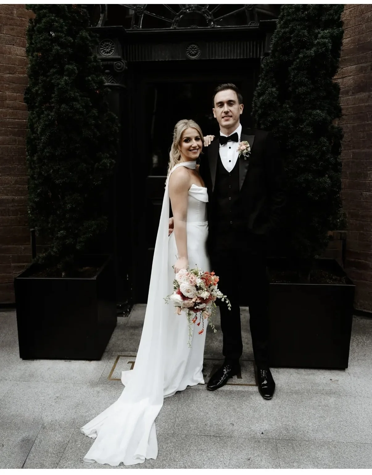 A bride and groom stand together in wedding attire, smiling for a photo outdoors in front of a brick building and black door with decorative black planters and greenery.