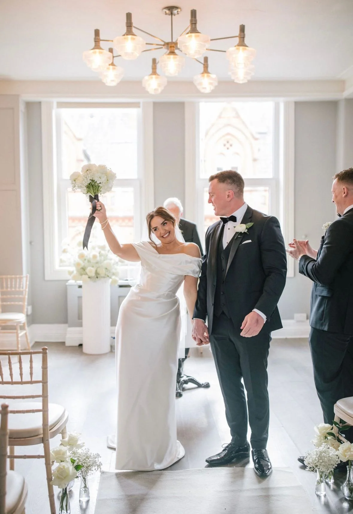 A bride in a white wedding dress holding a bouquet of white flowers in the air, smiling at the groom, who is dressed in a black tuxedo with a white shirt and black bow tie, in a bright, softly lit indoor space with large windows and floral decoration