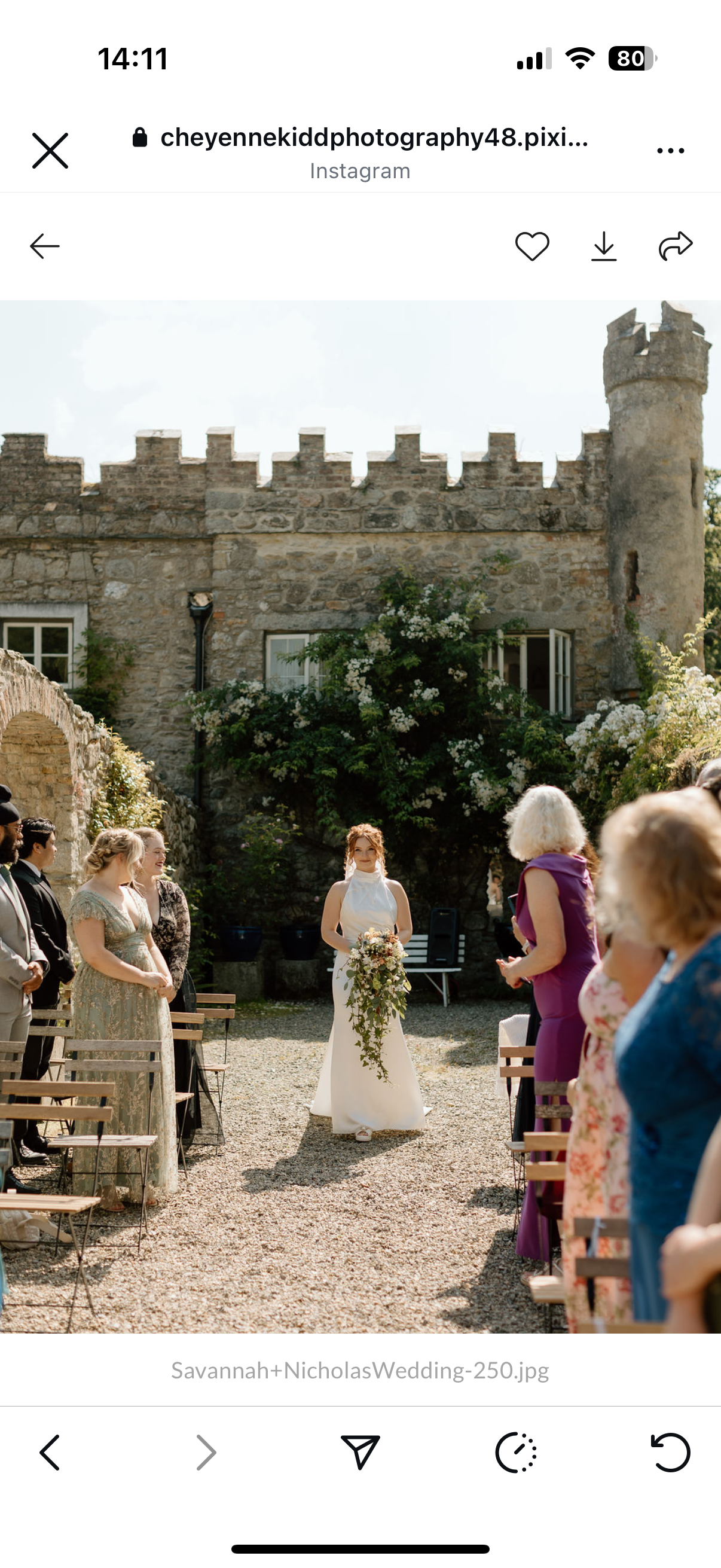 Bride walking down the aisle, holding a teat drop bouquet, at an outdoor wedding ceremony with guests on either side, in front of a stone castle wall with greenery.