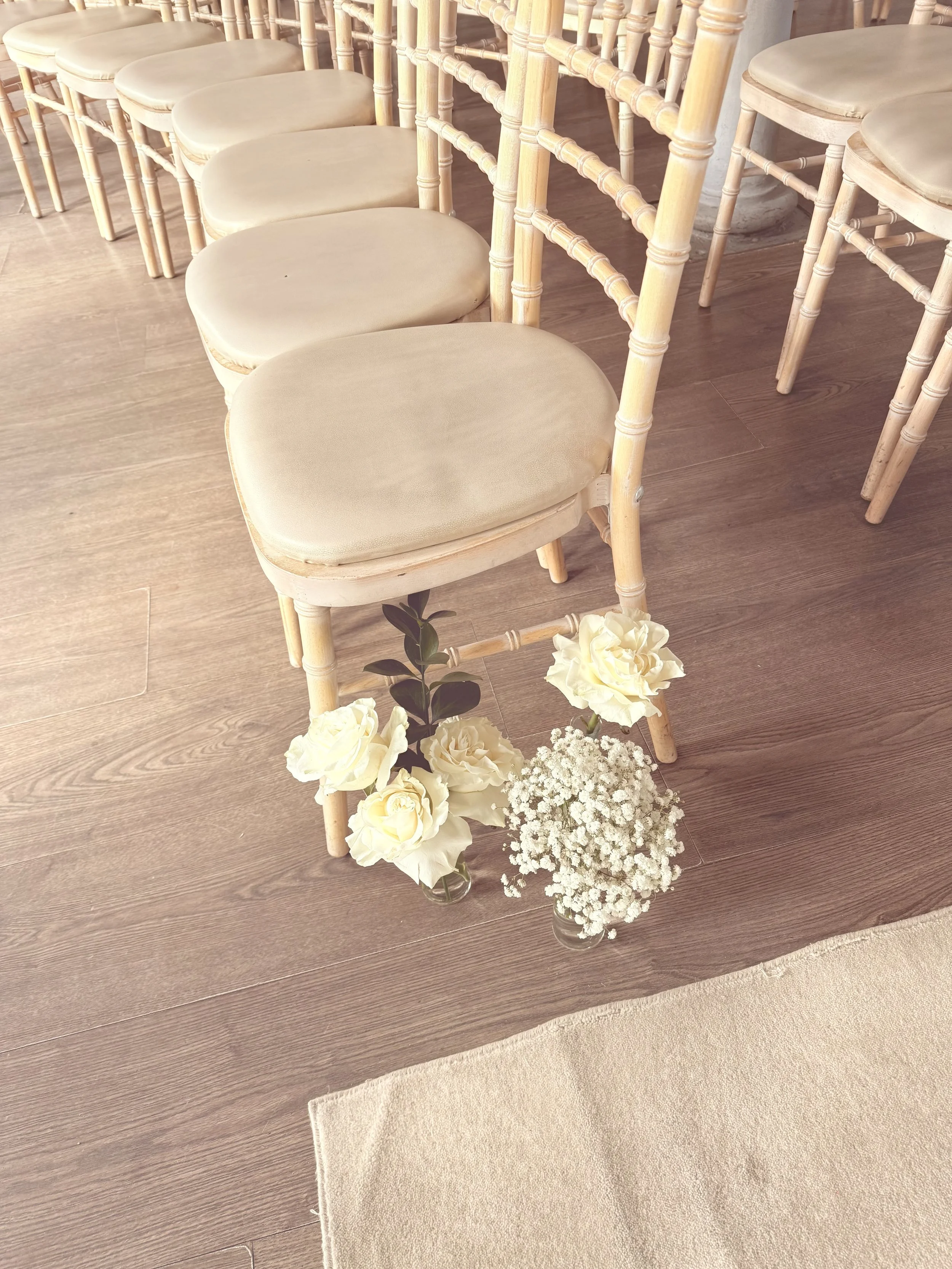 A row of beige cushioned chairs with wooden frames and a floral decoration at the base, consisting of white roses and baby's breath in glass vases, on a wooden floor near a beige rug.
