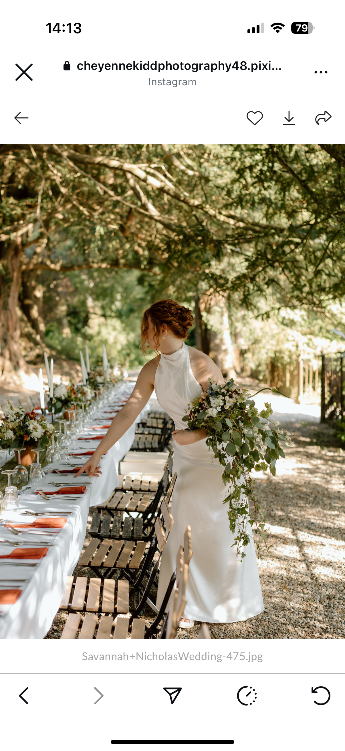 A bride in a white wedding dress adjusts a table setting outdoors under a large tree, holding a bouquet of flowers, with a long, decorated table set with plates, glasses, silverware, and floral arrangements.