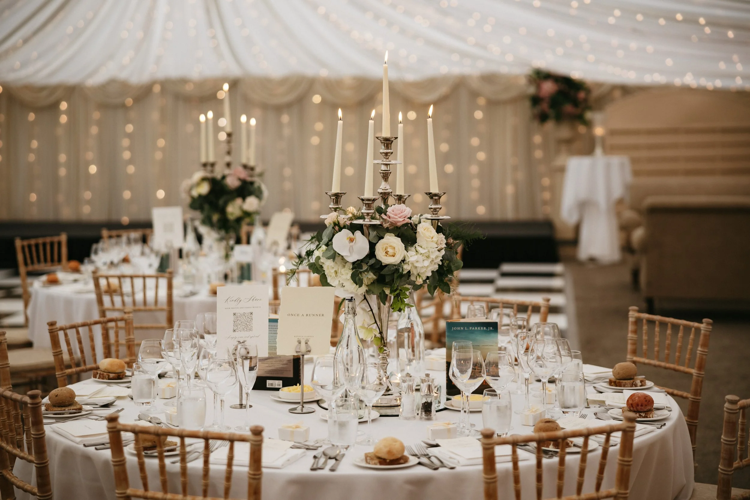 Elegant banquet table set for a wedding reception with a large floral centerpiece and tall white candles, dressed candelabras, surrounded by chairs and tableware, in a decorated venue with string lights and draped ceiling.