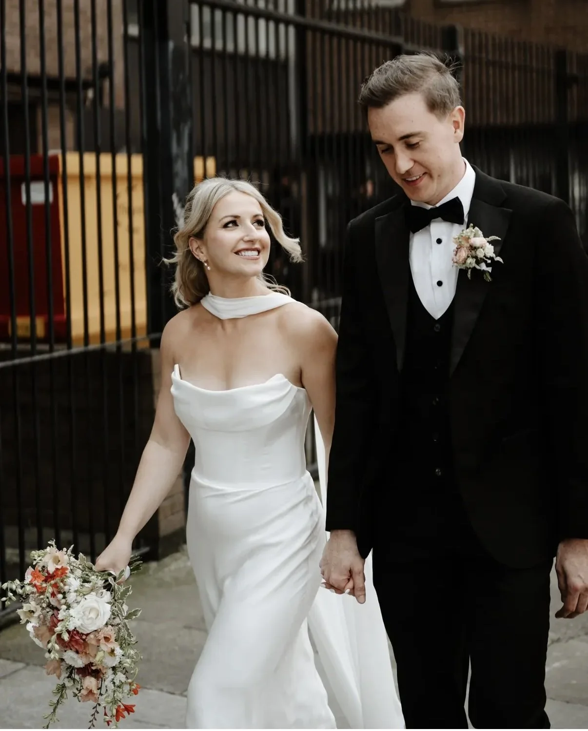 A bride and groom holding hands and walking outdoors, smiling at each other, with the bride holding a bouquet of flowers.
