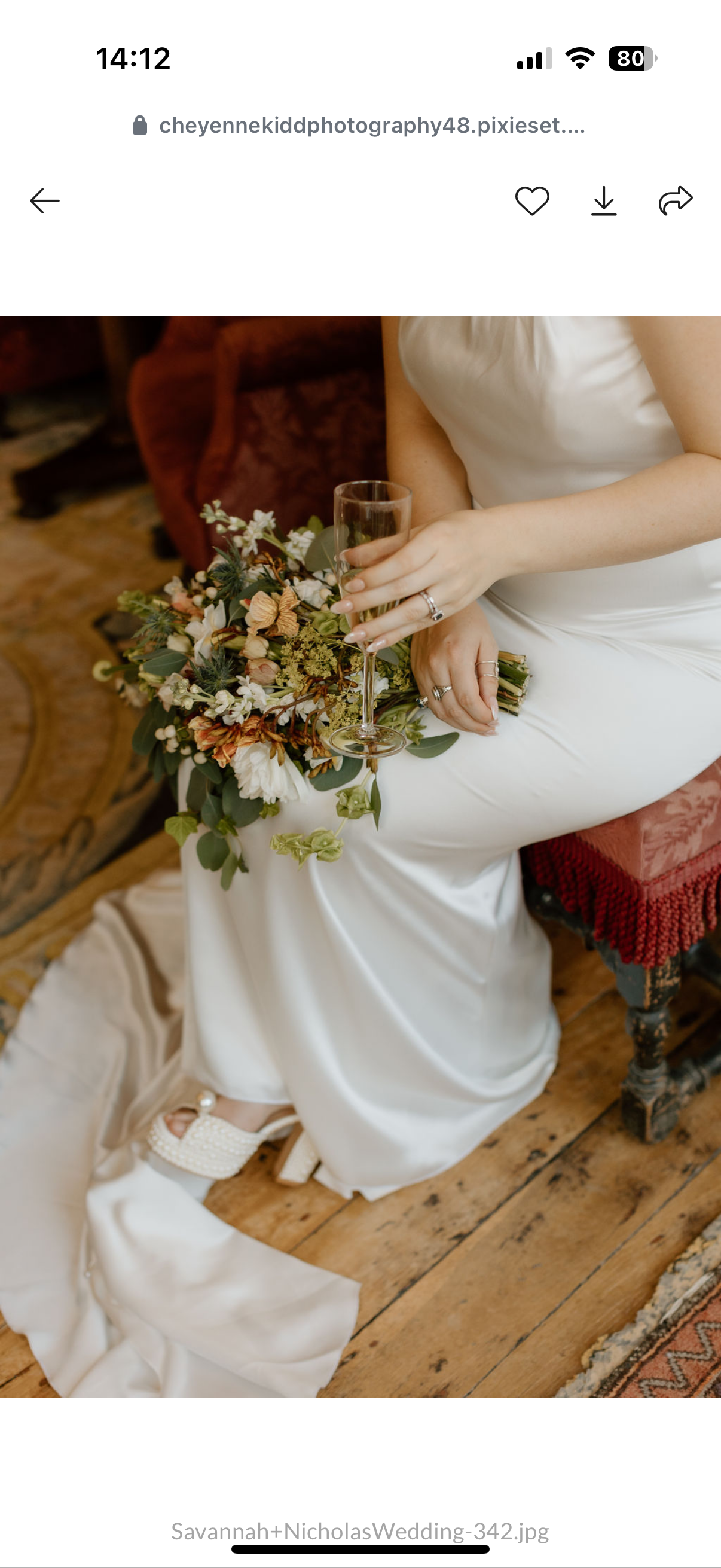 A woman in a white wedding dress sitting on a red velvet chair, holding a champagne glass, with a bouquet of flowers resting on her lap. Her left foot with a pearl shoe is visible on the wooden floor.