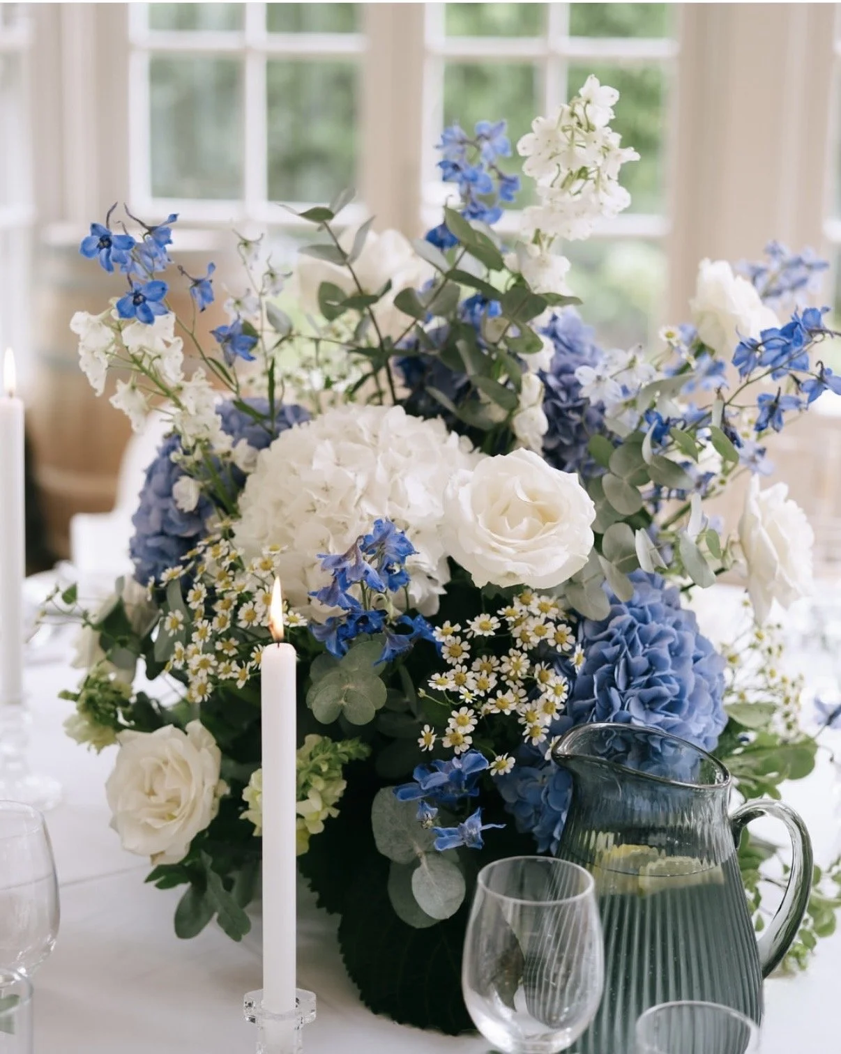 A floral centerpiece with white, blue, and green flowers on a white table, with a lit candle, a glass pitcher, and wine glasses, set against a window background.