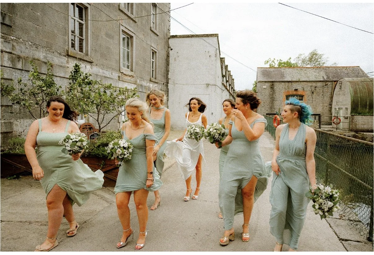 A group of seven women in pastel dresses walking outdoors, carrying bouquets of white flowers, with old buildings and trees in the background.