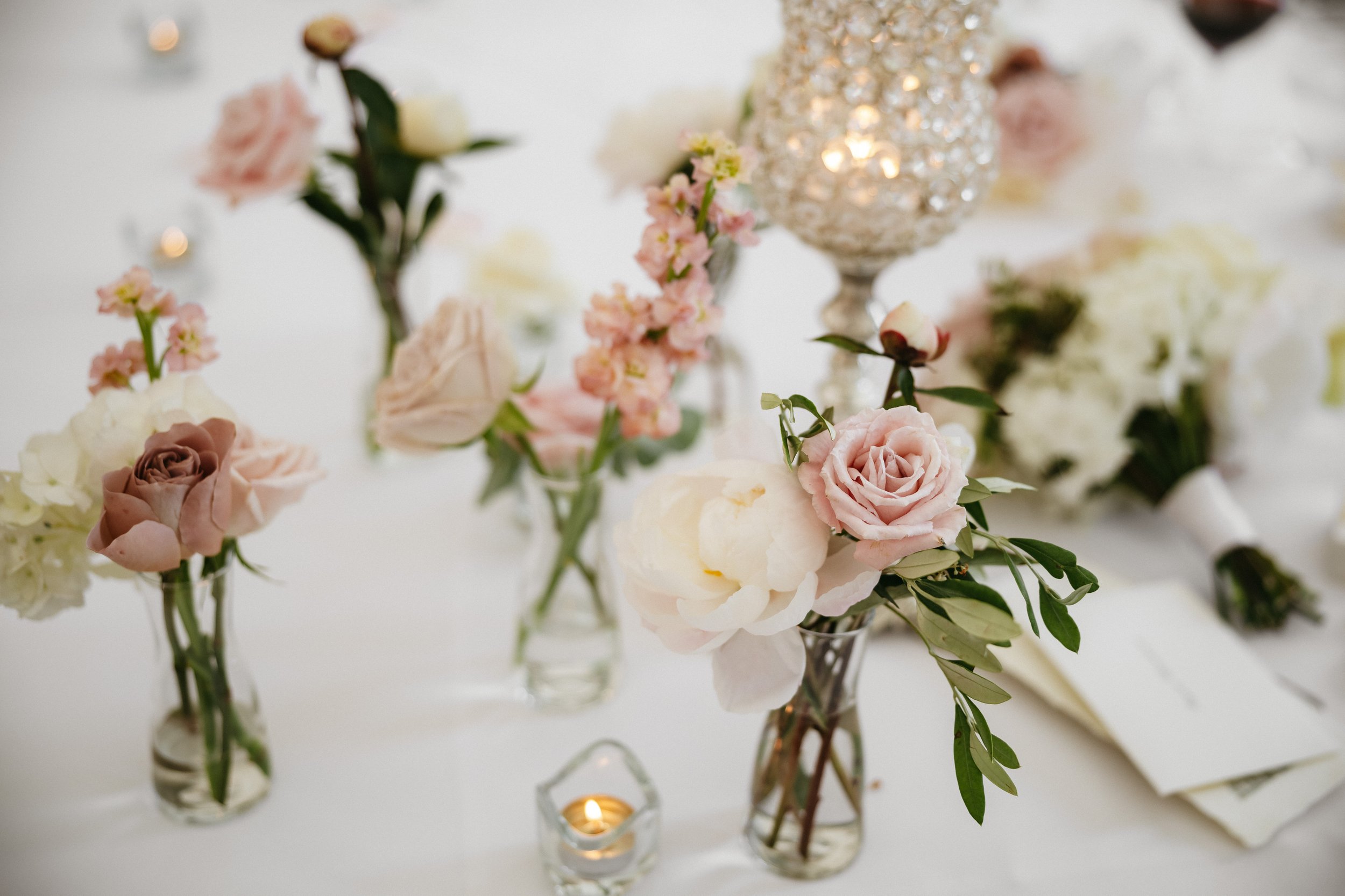 Elegant table centerpiece with pink and white flowers in small glass vases, bud vases, a lit candle in a glass holder, and a large crystal candle holder, all on a white tablecloth.
