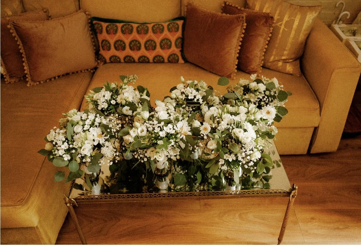 A floral arrangement with white flowers and green leaves on a glass-topped wooden coffee table in front of a yellow sofa with various decorative pillows.