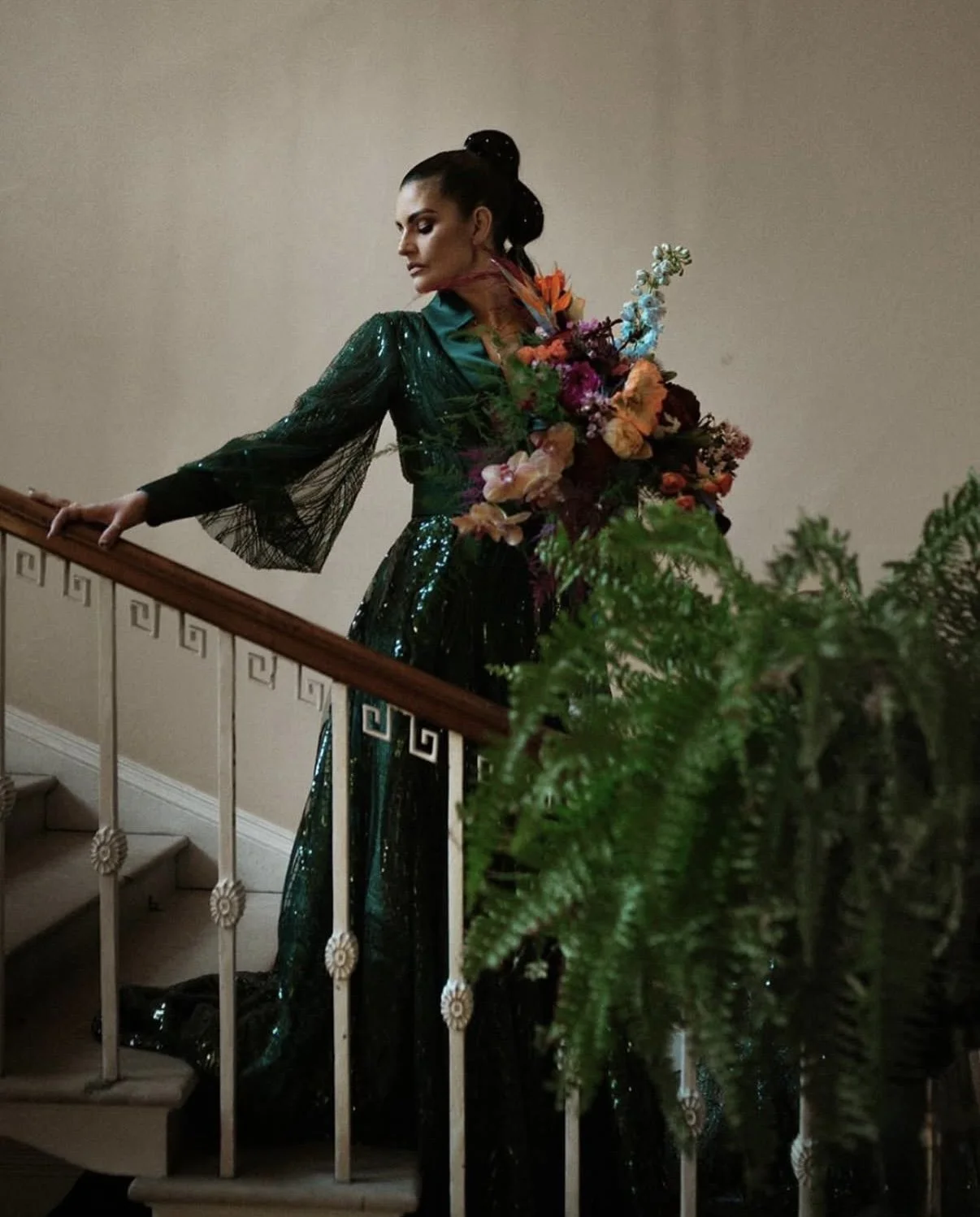 A woman in a dark green, shiny evening gown with sheer black sleeves stands on a staircase, holding a large bouquet of colorful flowers on her back, near a potted fern plant.