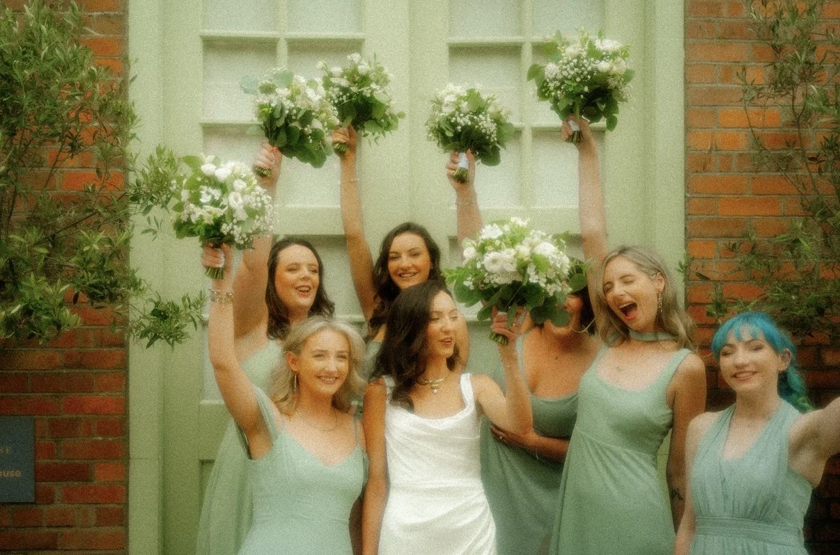 Group of women celebrating, holding bouquets, in front of green door with brick walls, wearing pastel dresses, joyful and smiling.