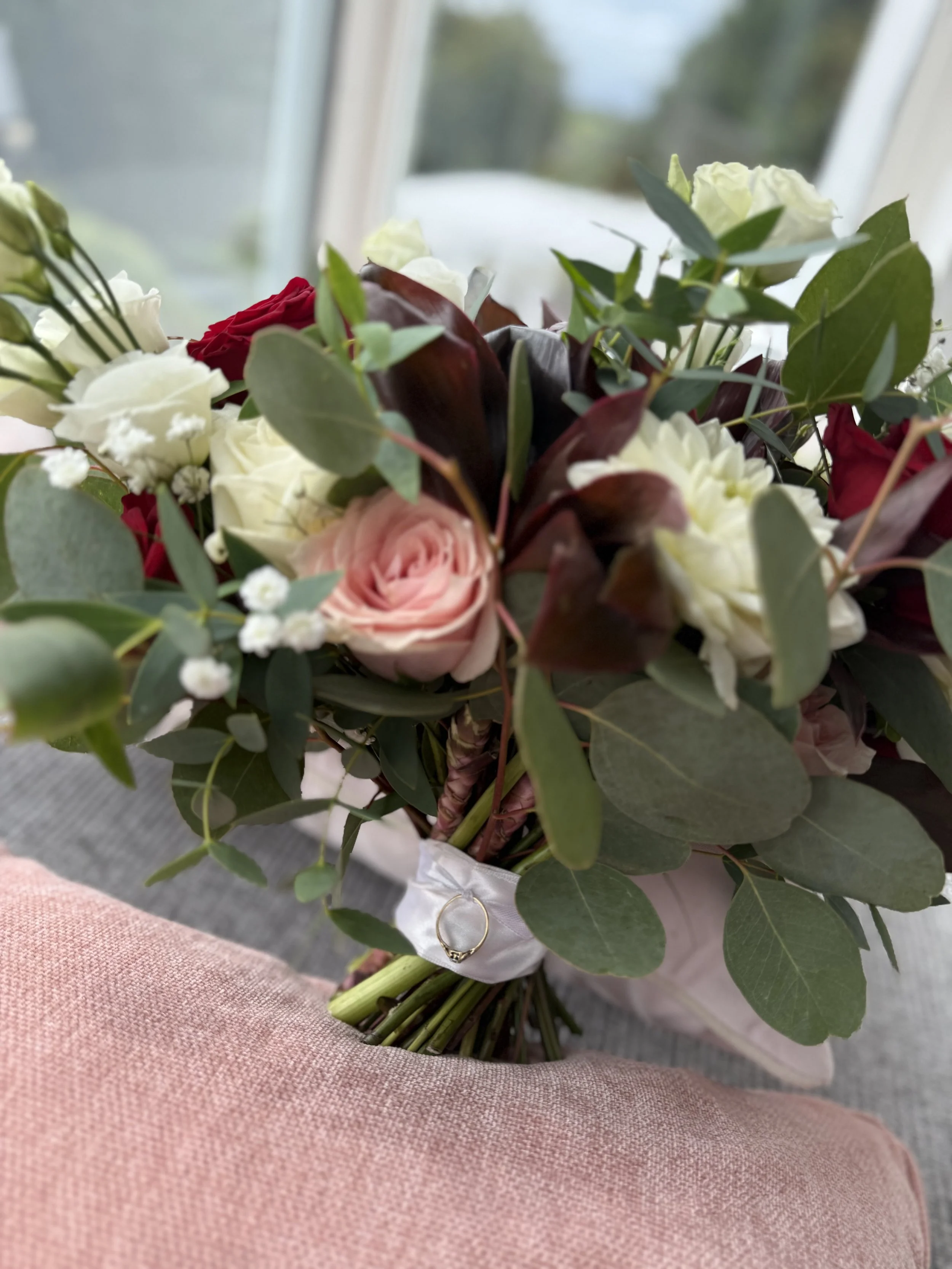 A bouquet of roses, greenery, and decorative flowers wrapped with a white ribbon, resting on a pink surface near a window.