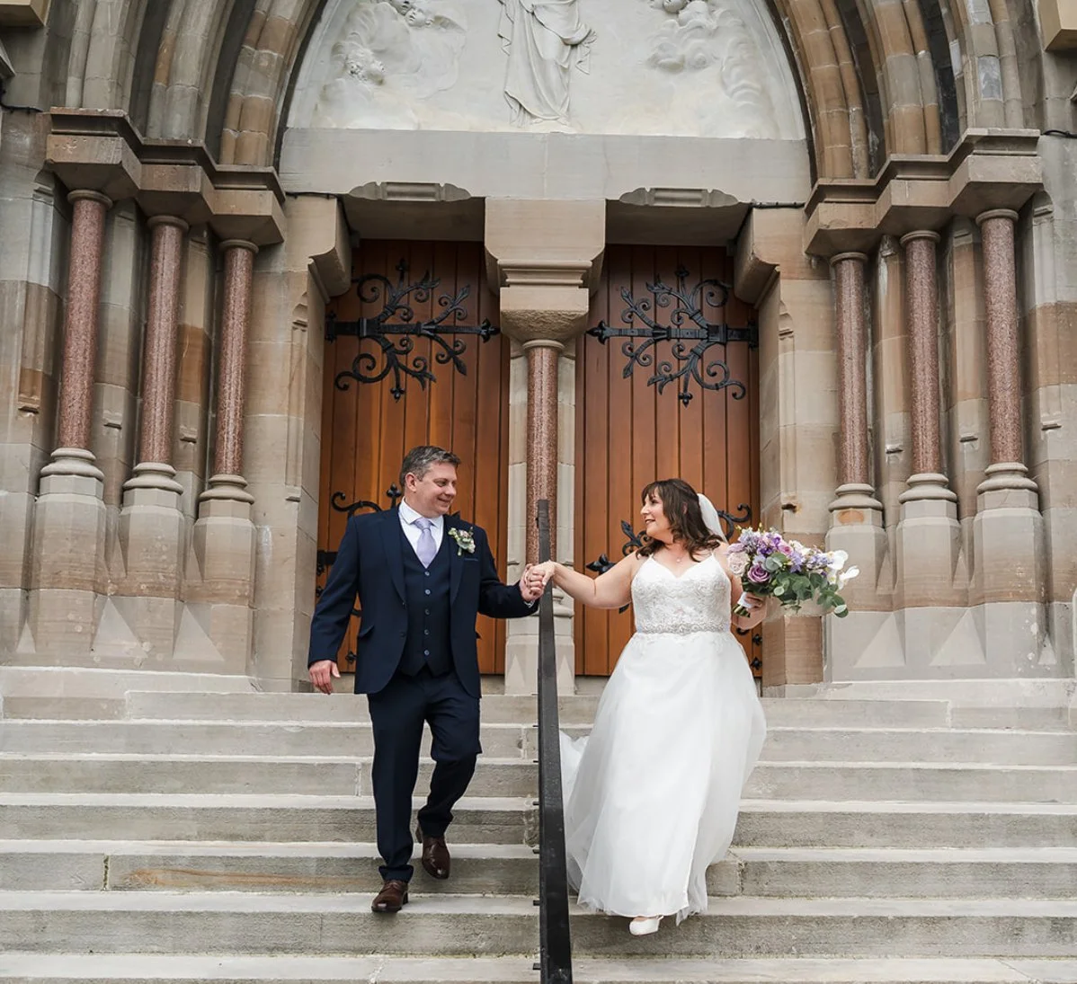 A bride and groom holding hands on the stairs in front of a church entrance, with ornate wooden doors and stone columns, smiling at each other. The bride is carrying a bouquet of flowers.