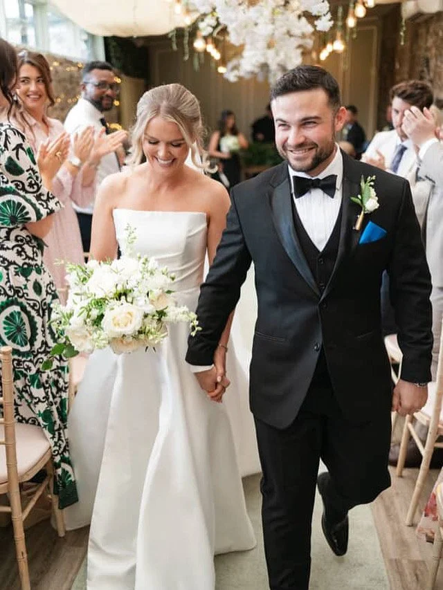 A newlywed couple holding hands and smiling at their wedding reception, surrounded by friends, with floral decorations and warm lighting.