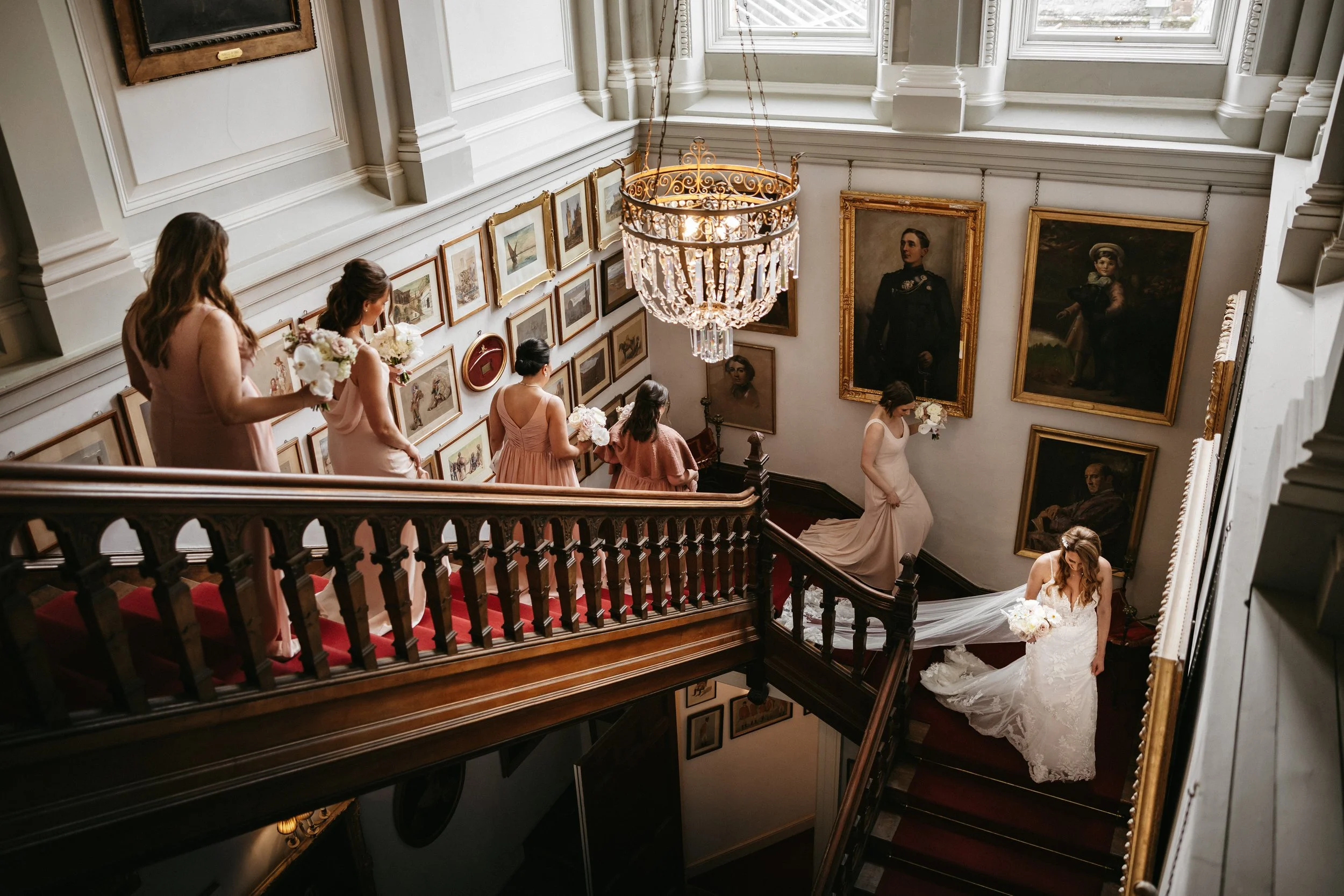 A bride in a white lace wedding gown with a train holds a bouquet as she walks up a grand staircase inside a museum or art gallery. Five bridesmaids in pink dresses follow her, each holding a bouquet. The staircase has red carpet, and the walls are d