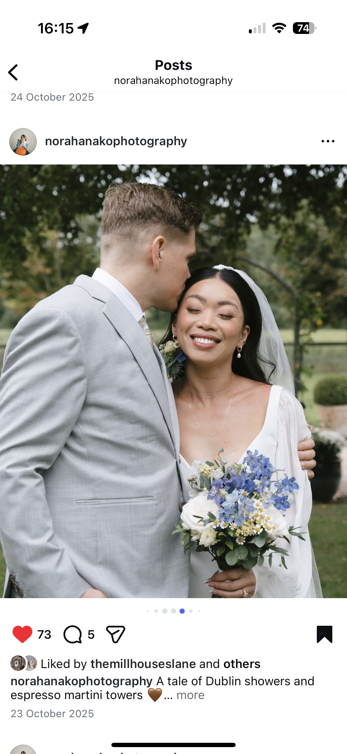 A bride and groom embracing outdoors, with the groom kissing the bride's forehead. The bride has dark hair, is smiling, and holding a bouquet of blue and white flowers. The groom is wearing a light gray suit, and the background features greenery.
